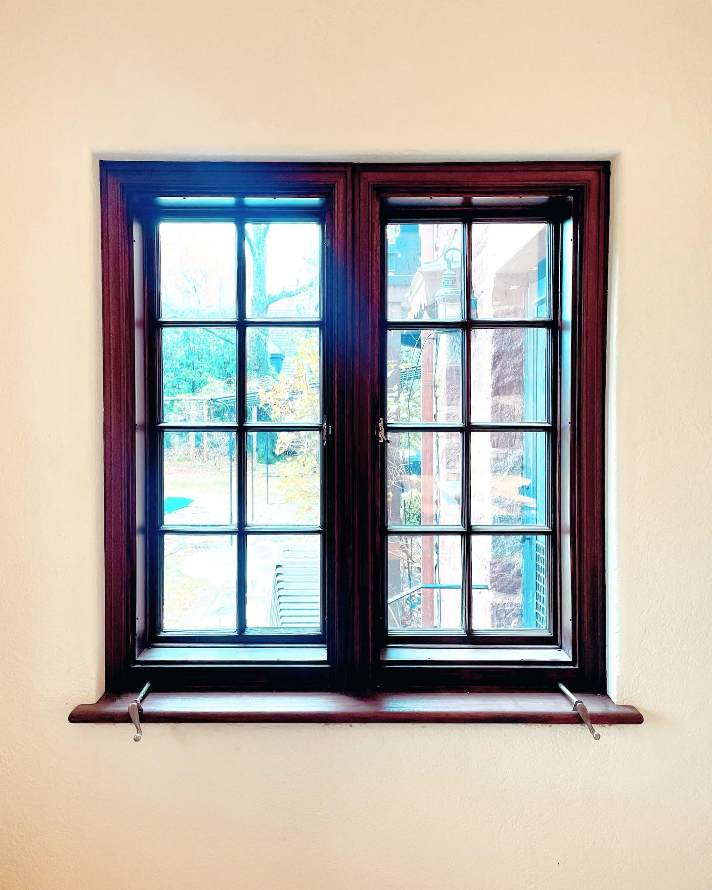 A double-pane window with dark wood trim set in a light-colored wall, with a view of an outdoor garden and neighboring building. Heartwood Renovations. West Hartford, CT
