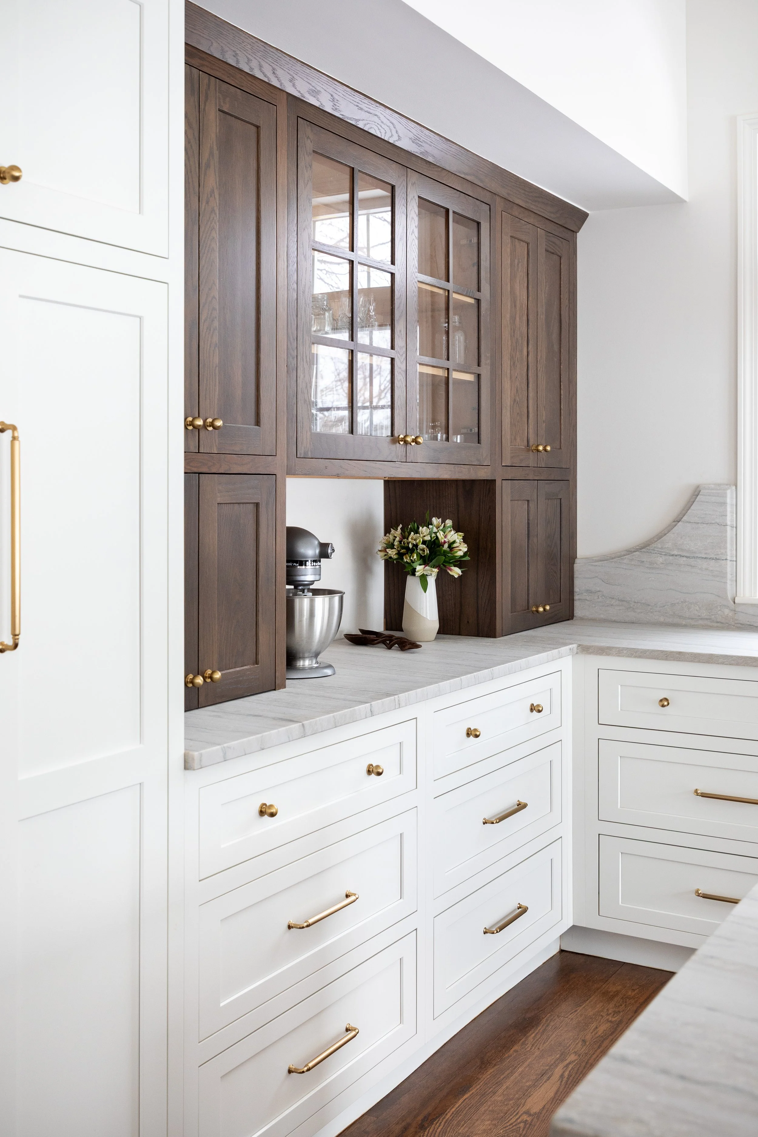 Kitchen cabinetry with white lower cabinets and dark brown upper cabinets, a marble countertop, a stand mixer, a vase of flowers, and a window reflecting outside scenery. Heartwood Renovations. West Hartford, CT