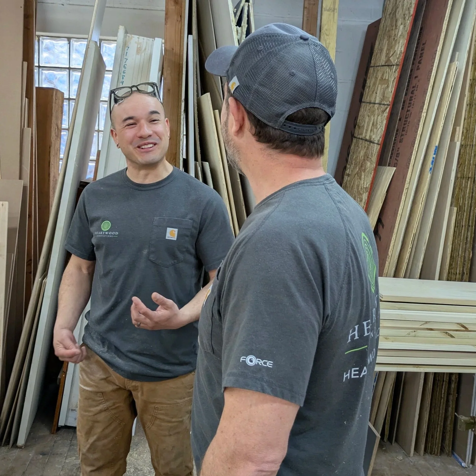 Marcos and Mike, Heartwood carpenters, talking in the shop beside stacked trim and millwork