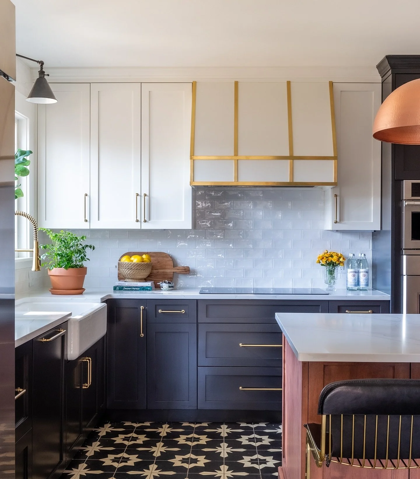 Modern kitchen with white upper cabinets, black lower cabinets with gold handles, a white countertop, a white subway tile backsplash, and decorative items including a green plant, a basket of lemons, and bottled water. Heartwood Renovations.