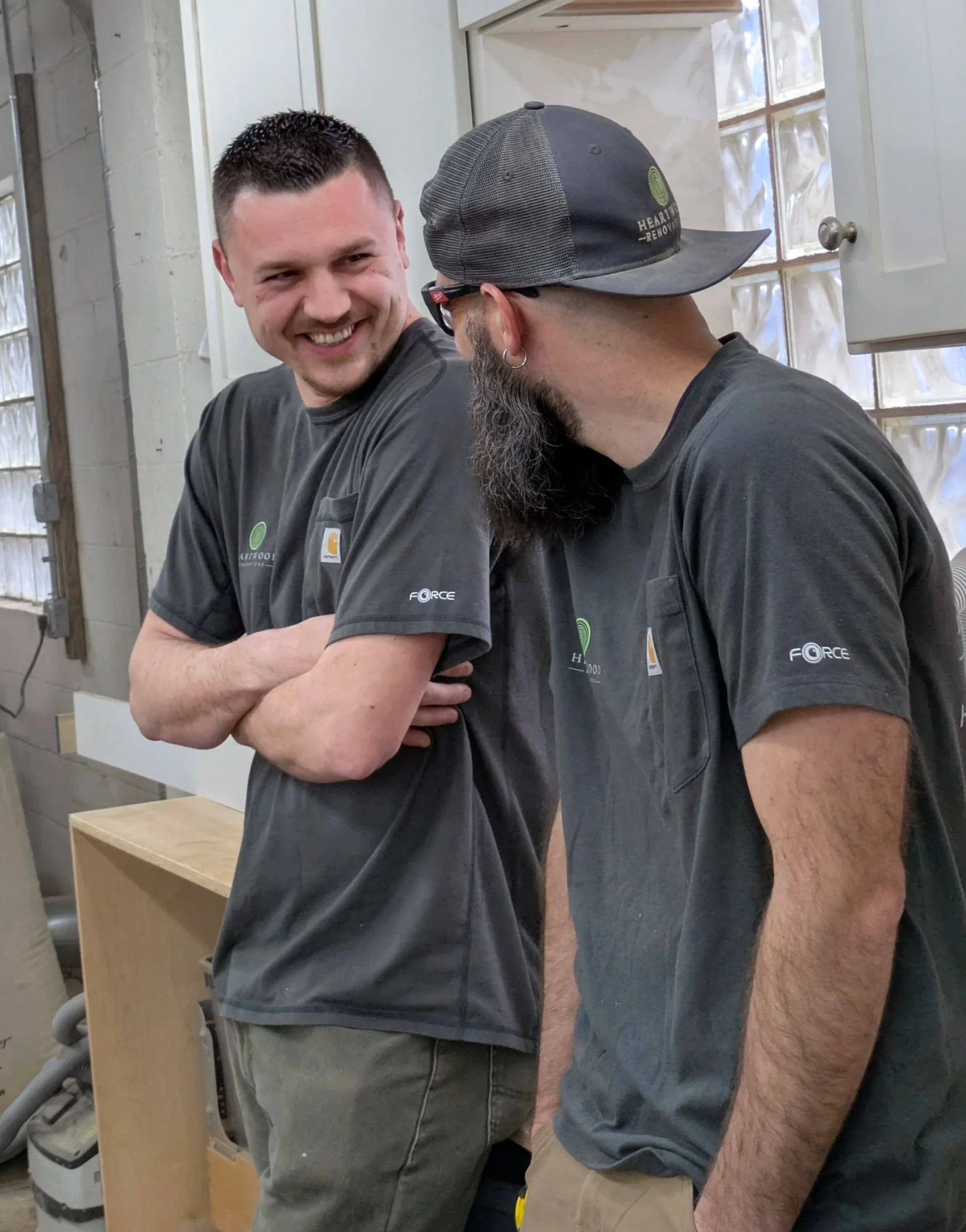 Two men with gray shirts, tattoos and facial hair having a friendly conversation in a workshop or garage. One man is smiling and looking at the other, who is wearing glasses, a baseball cap, and earrings. Heartwood Renovations CT