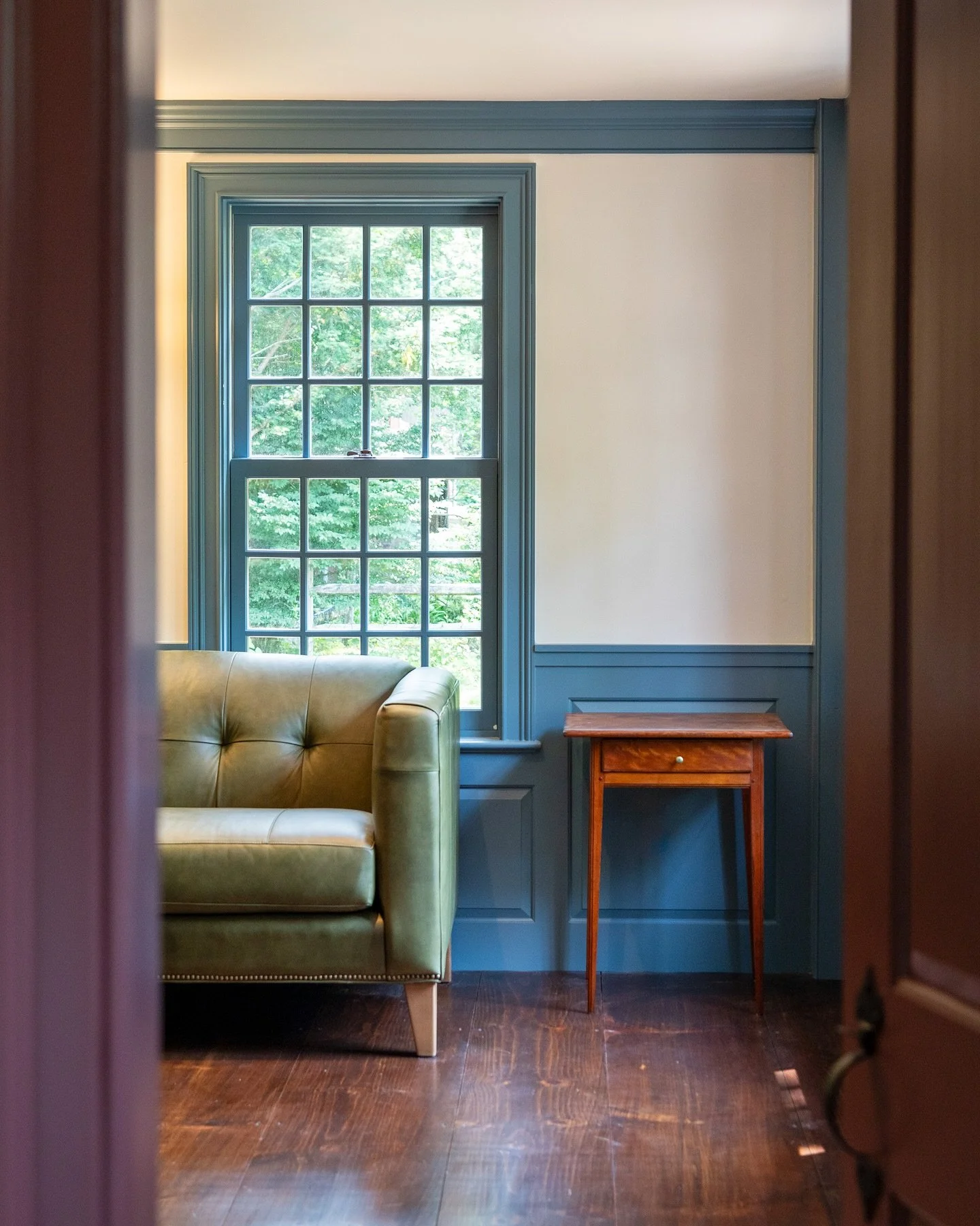 A cozy room with a large window, a green leather armchair, and a wooden side table with a drawer. Heartwood Renovations. West Hartford, CT