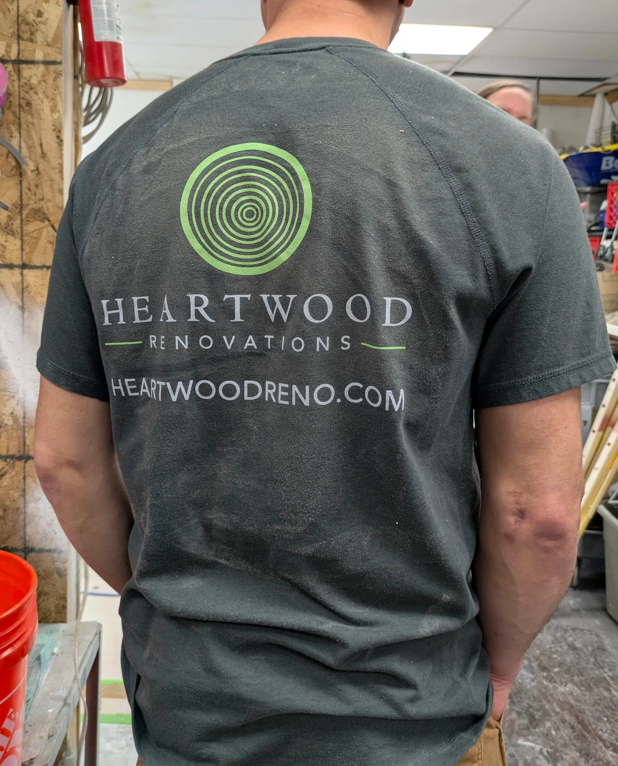 Back of a person wearing a black T-shirt with green circular logo and text for Heartwood Renovations, including the website heartwoodreno.com, in a workshop or hardware store. Heartwood Renovations. West Hartford, CT