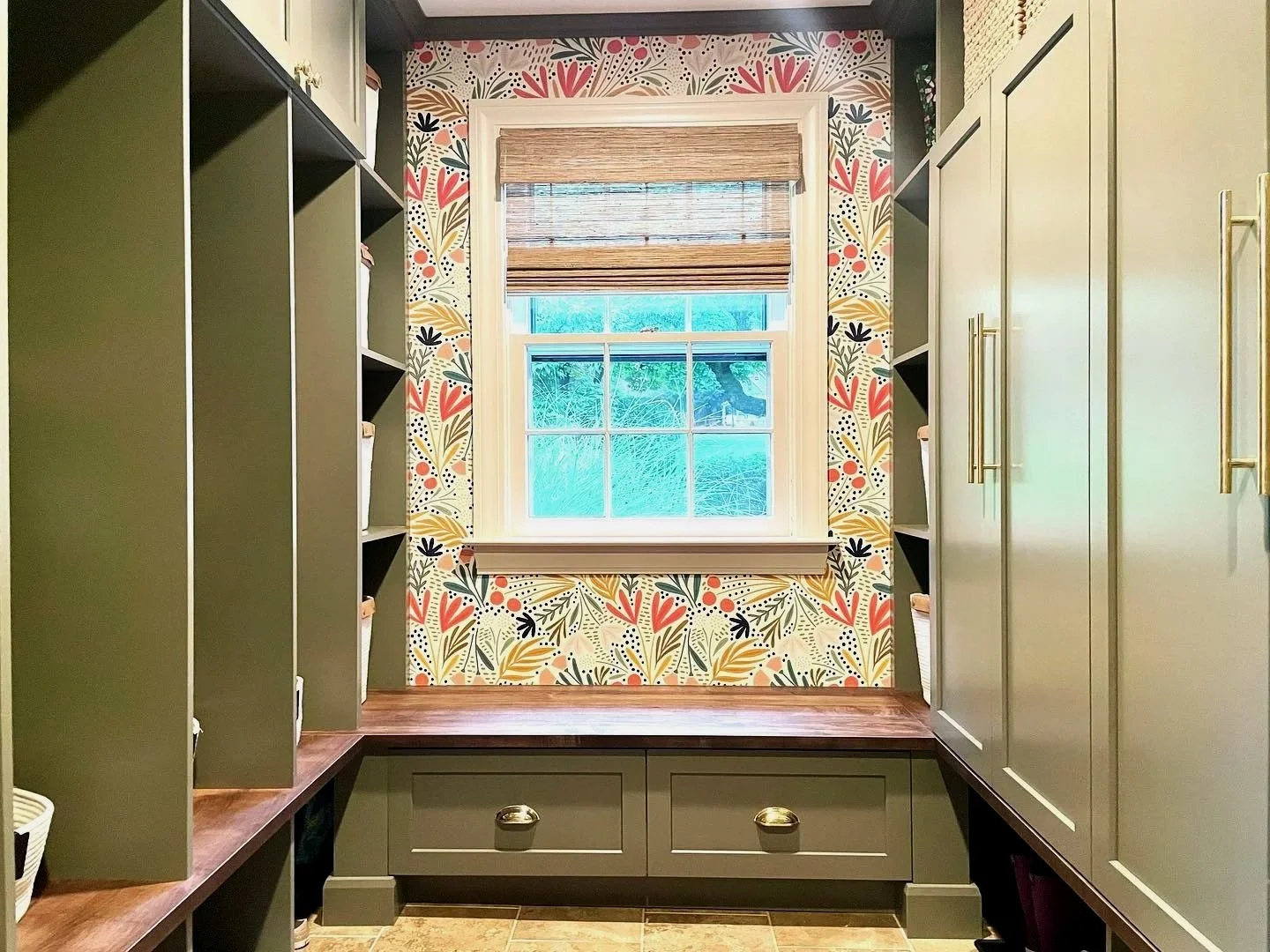 A walk-in closet with built-in shelves and cabinets on both sides, a window with a bamboo shade in the center, floral-patterned wallpaper around the window, and a wooden bench with drawers below the window. Heartwood Renovations. West Hartford, CT