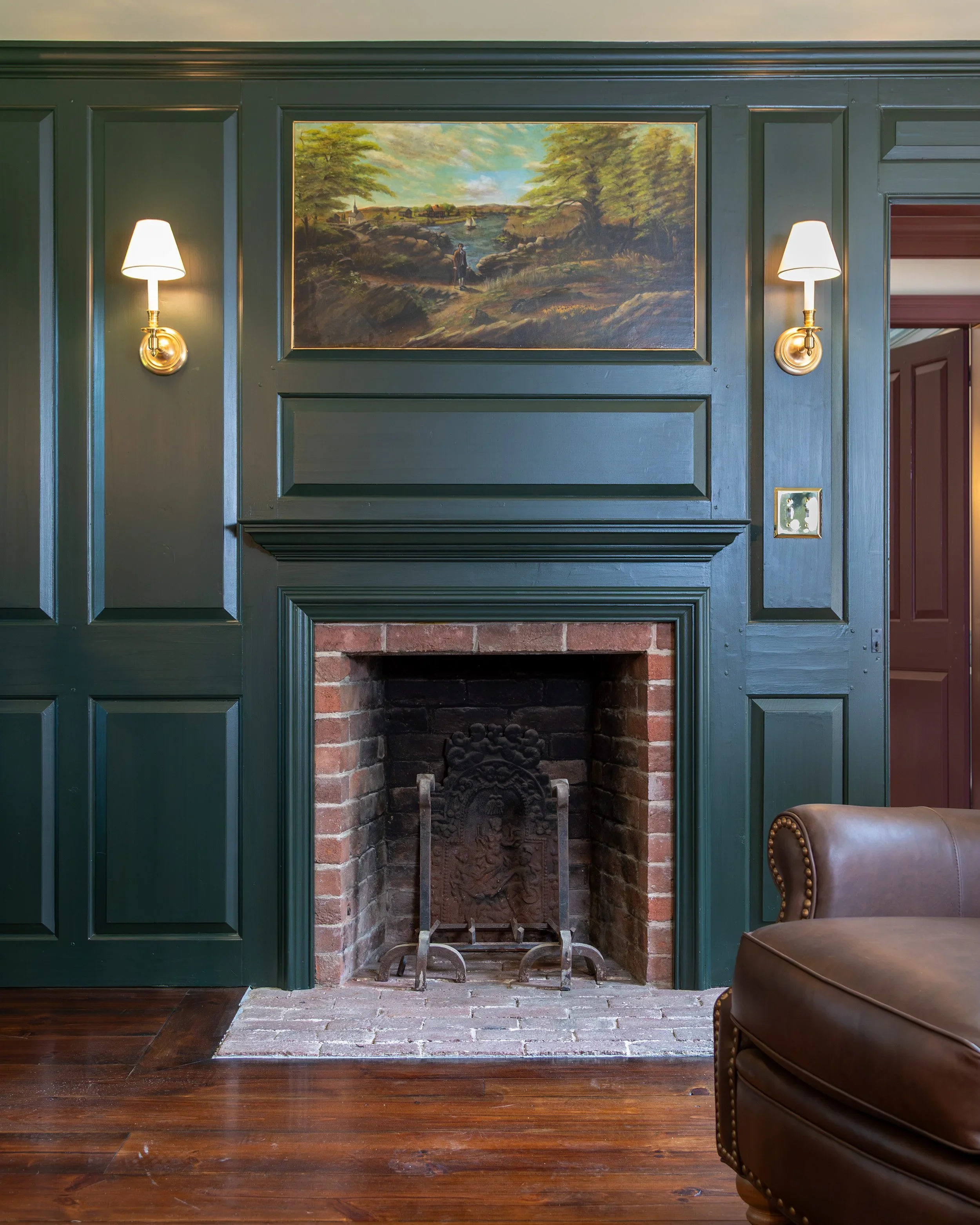 A fireplace with a dark green wooden mantel and paneling, a painting above it depicting a landscape with trees and a person, flanked by two wall lamps with white shades, a brown leather chair on the right, and hardwood flooring. Heartwood Renovations