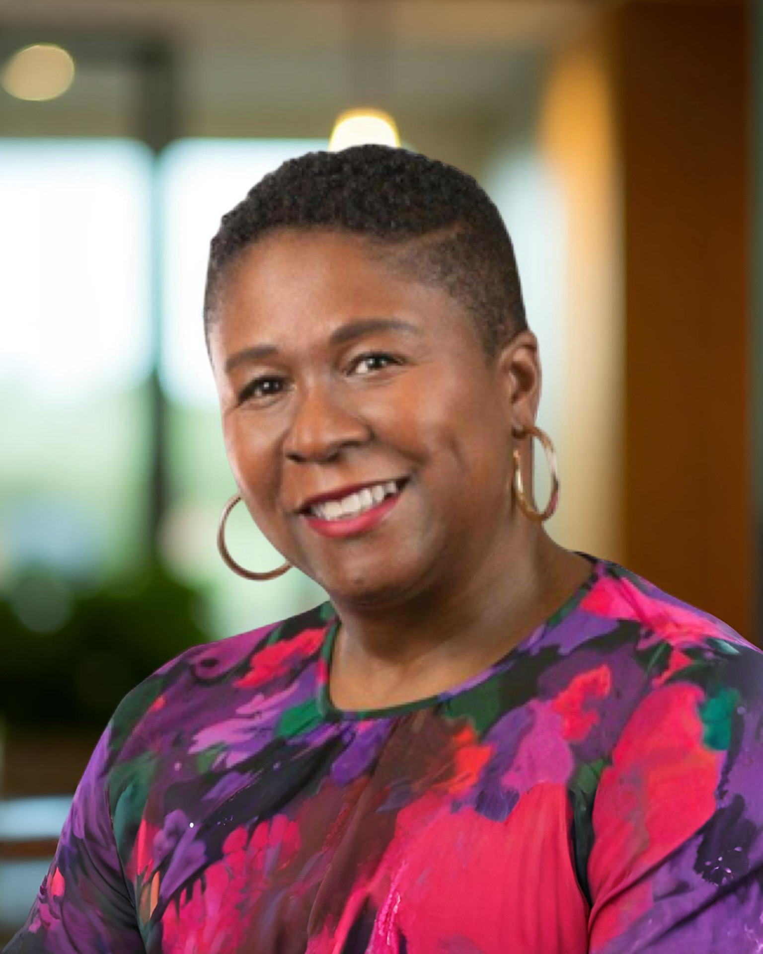 Portrait of a smiling African American woman with short curly hair, wearing hoop earrings and a colorful purple, pink, and green floral blouse, in a bright indoor setting.