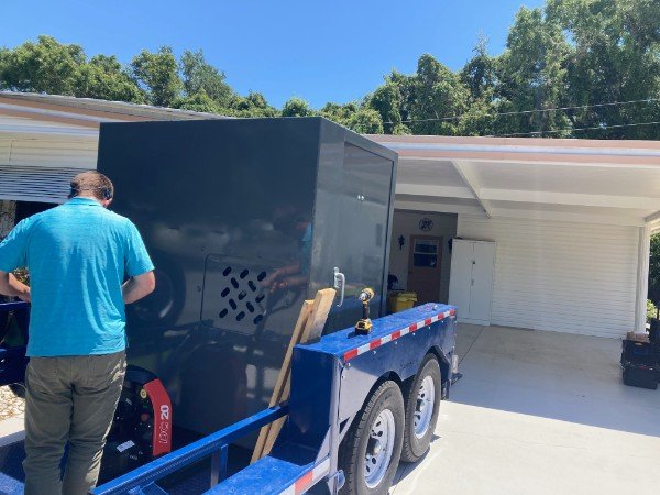 A man in a blue shirt and gray pants standing next to a large black box mounted on a blue trailer in a driveway, with a house and garage in the background.