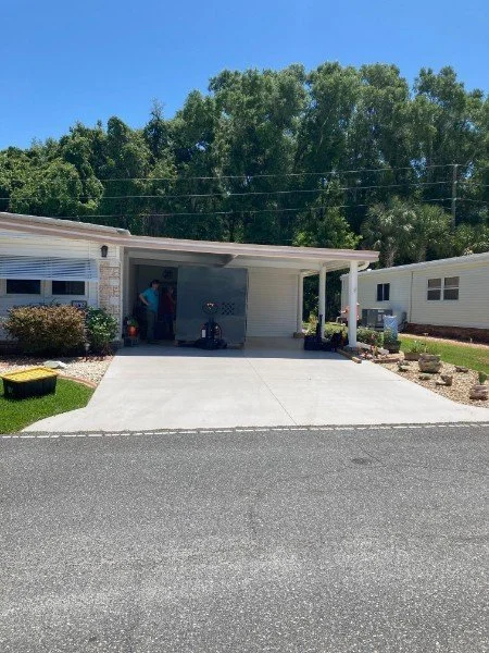 View of a residential house with a carport, a driveway, and some yard decorations. A person is standing inside the garage. Trees and blue sky in the background.