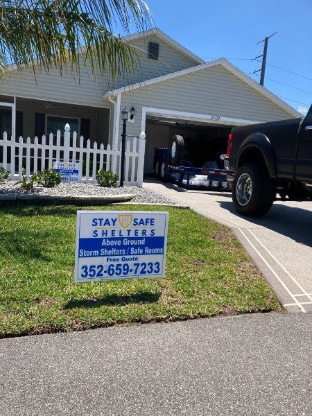 A house with a white picket fence and a sign in the yard advertising stay safe shelters, storm shelters, and safe rooms with a phone number. A black truck is parked in the driveway.
