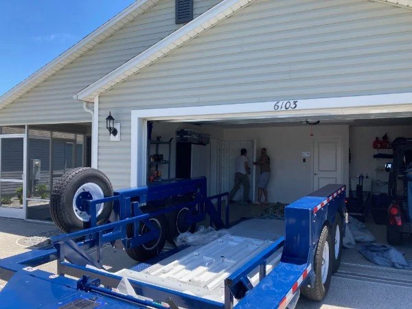 Blue flatbed trailer parked in front of a house garage with a white exterior, with two people inside the garage.