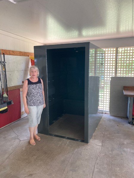 Older woman standing next to a large empty black metal box in a workshop with tools and wooden wall paneling.
