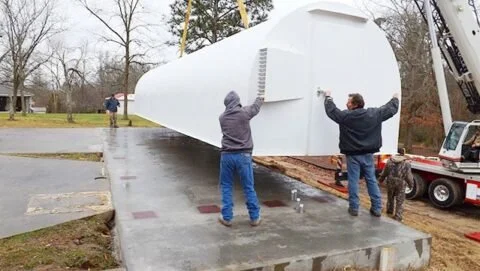 Two men guiding a large white industrial tank onto a trailer with a dog nearby, in an outdoor parking lot while rain falls.