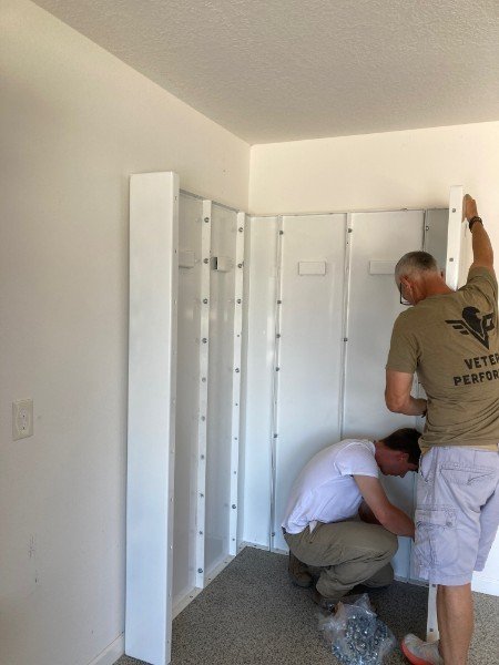 Two men assembling a white, folding partition wall inside a room with white walls and carpeted floor.