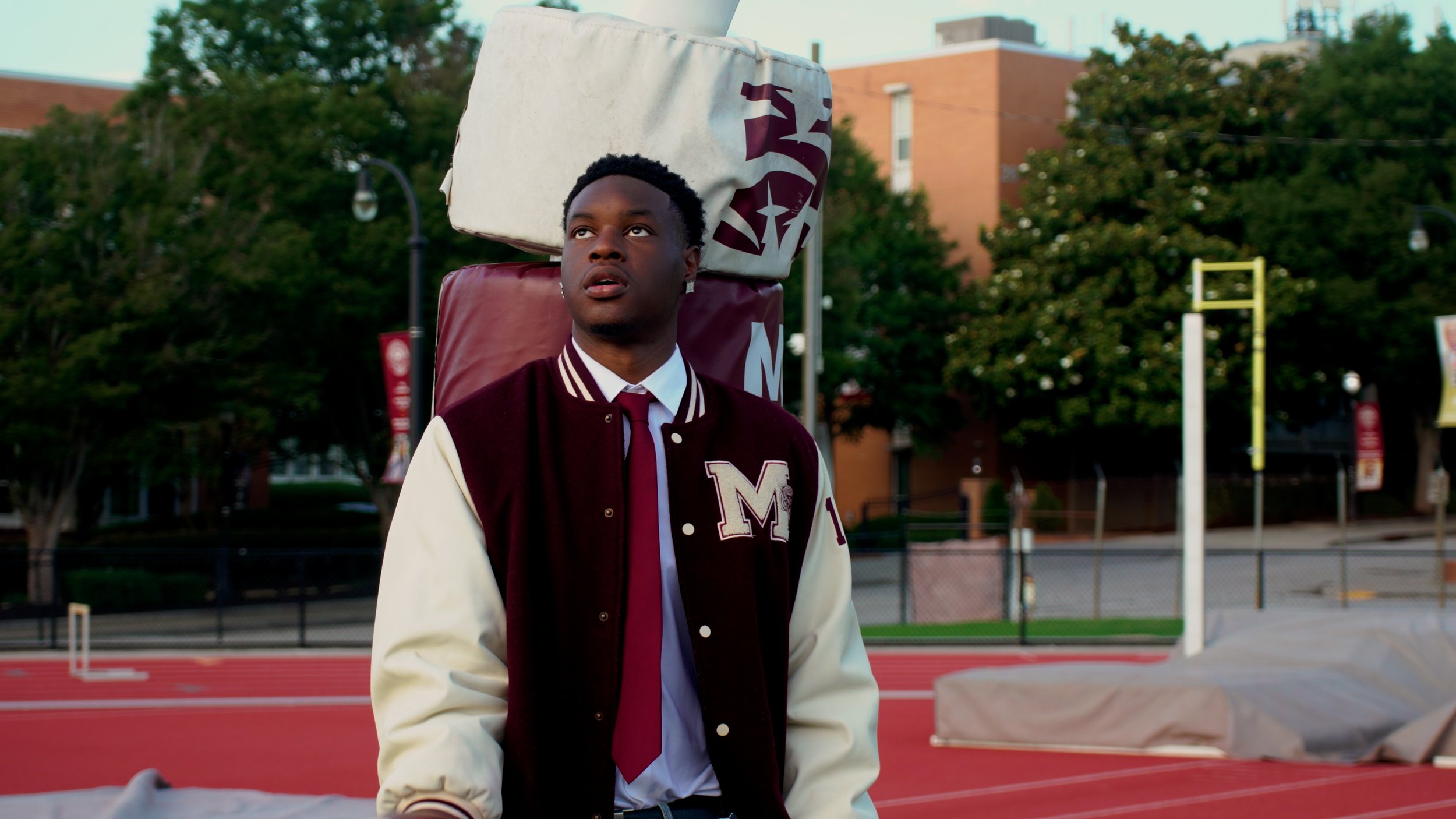 A young man with dark skin and short curly hair standing on a school track field. He is wearing a maroon and white letterman jacket with the letter 'M' on it, a white shirt, and a maroon tie. He has a large bag or padding strapped behind his shoulders. In the background, there are trees, a chain-link fence, and a brick building.