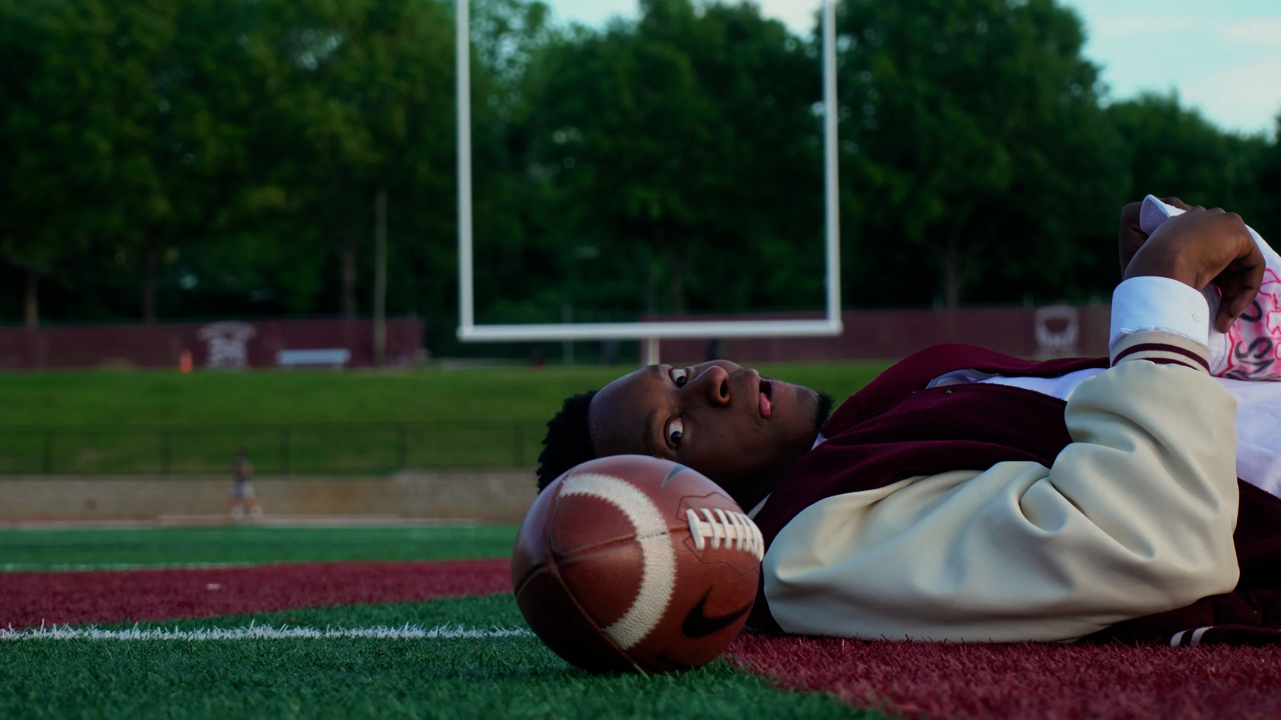 A young man lies on a football field, resting his head on a football while looking towards the camera, with football goalposts and trees in the background.