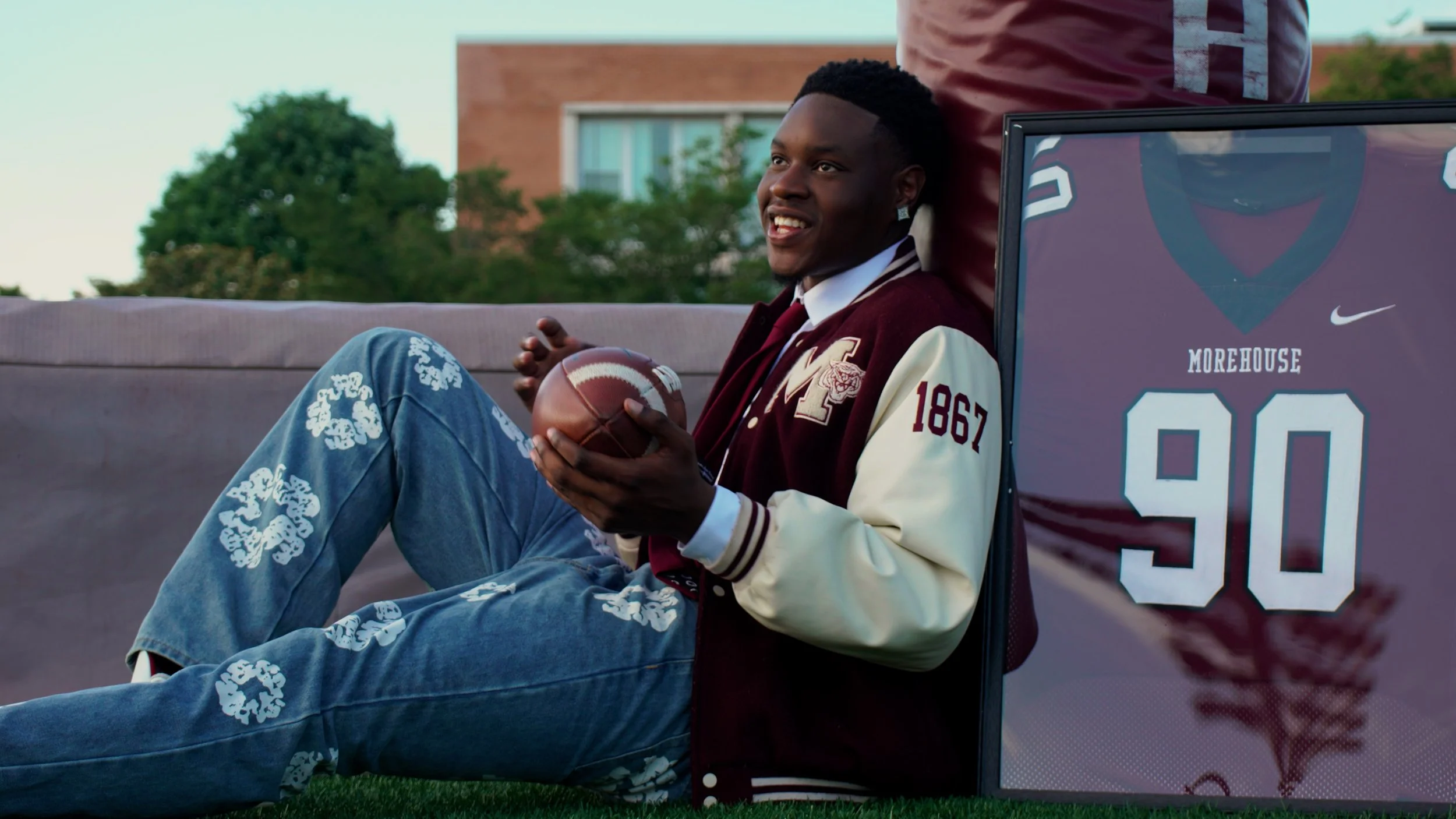 A young man sitting outdoors on a bench, holding a football, smiling, with a framed sports jersey with the name 'Morehouse' and number 90 beside him.