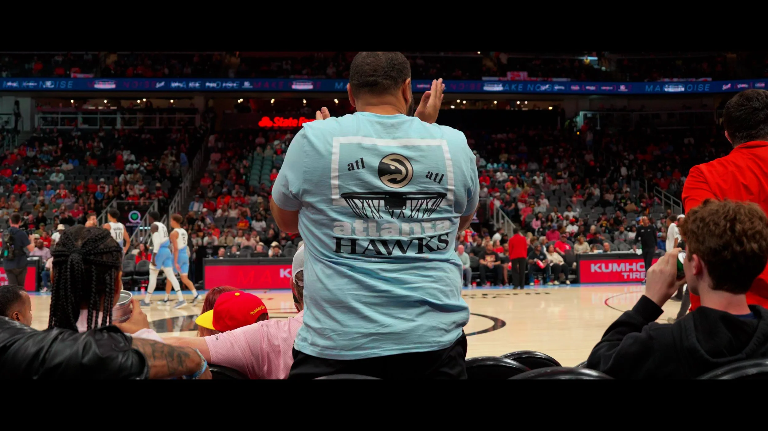 A man seated in an arena, wearing a light blue Atlanta Hawks T-shirt, watching a basketball game. The arena is filled with spectators, and the court is visible with players and digital advertisements.