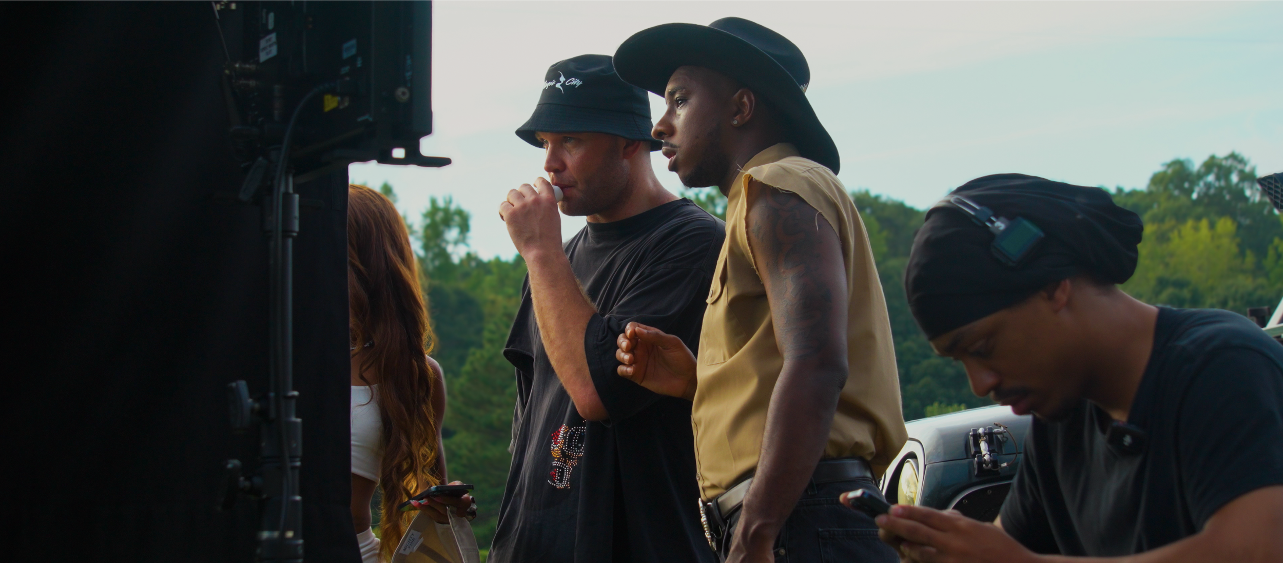 Group of people on set, filming outdoors with trees in background, two individuals looking at a monitor and one looking at a phone.