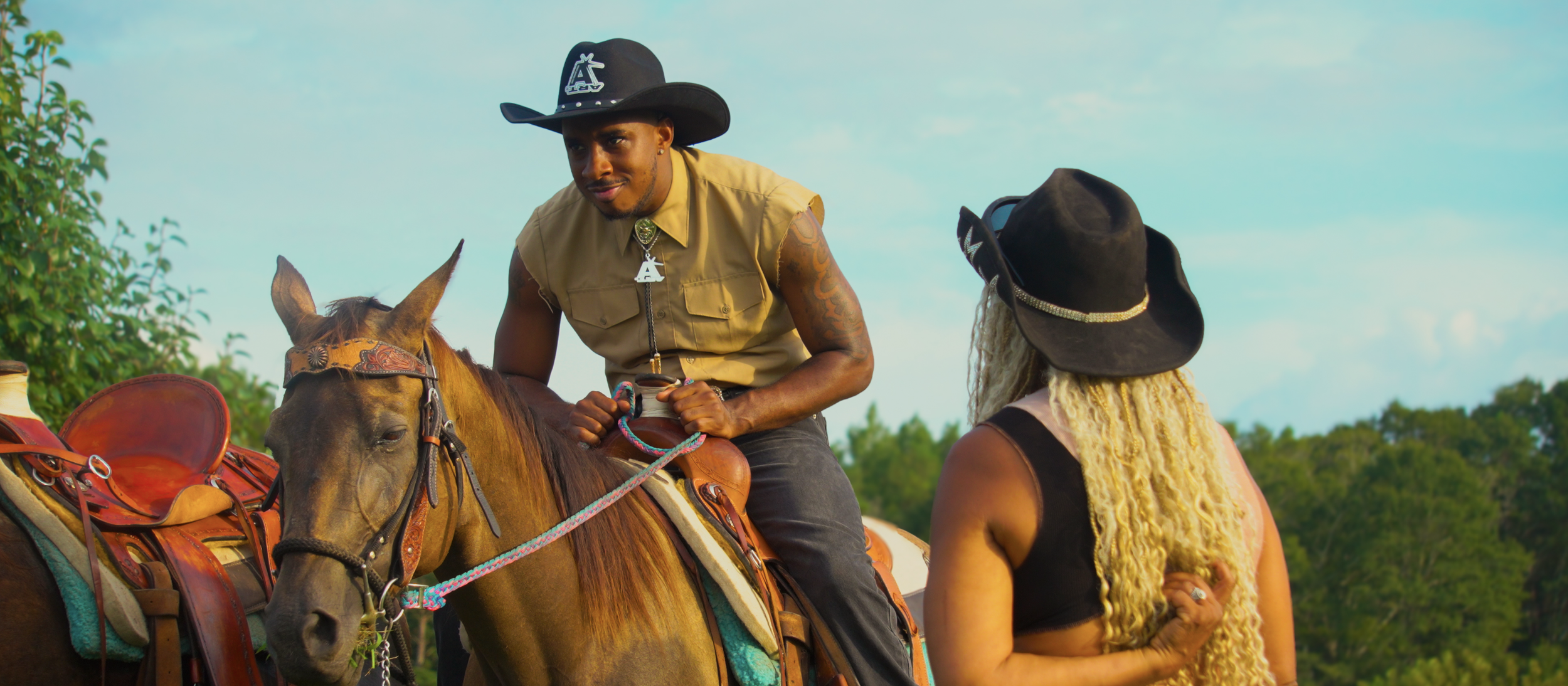 A man with tattoos wearing a cowboy hat and sleeveless shirt sits on a horse, talking to a woman with long blonde curly hair, wearing a black hat and sleeveless top, outdoors with trees and a blue sky in the background.