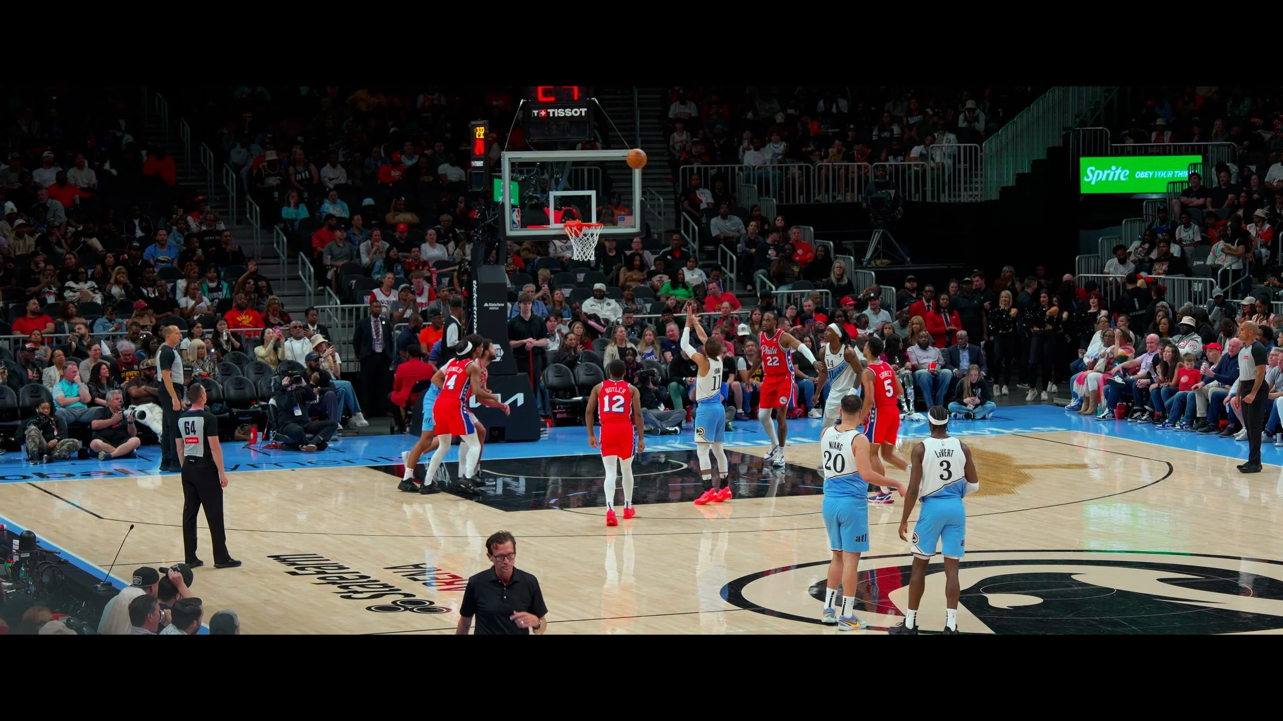 A basketball game taking place in an indoor arena with players in red and light blue uniforms on the court, surrounded by fans and photographers.