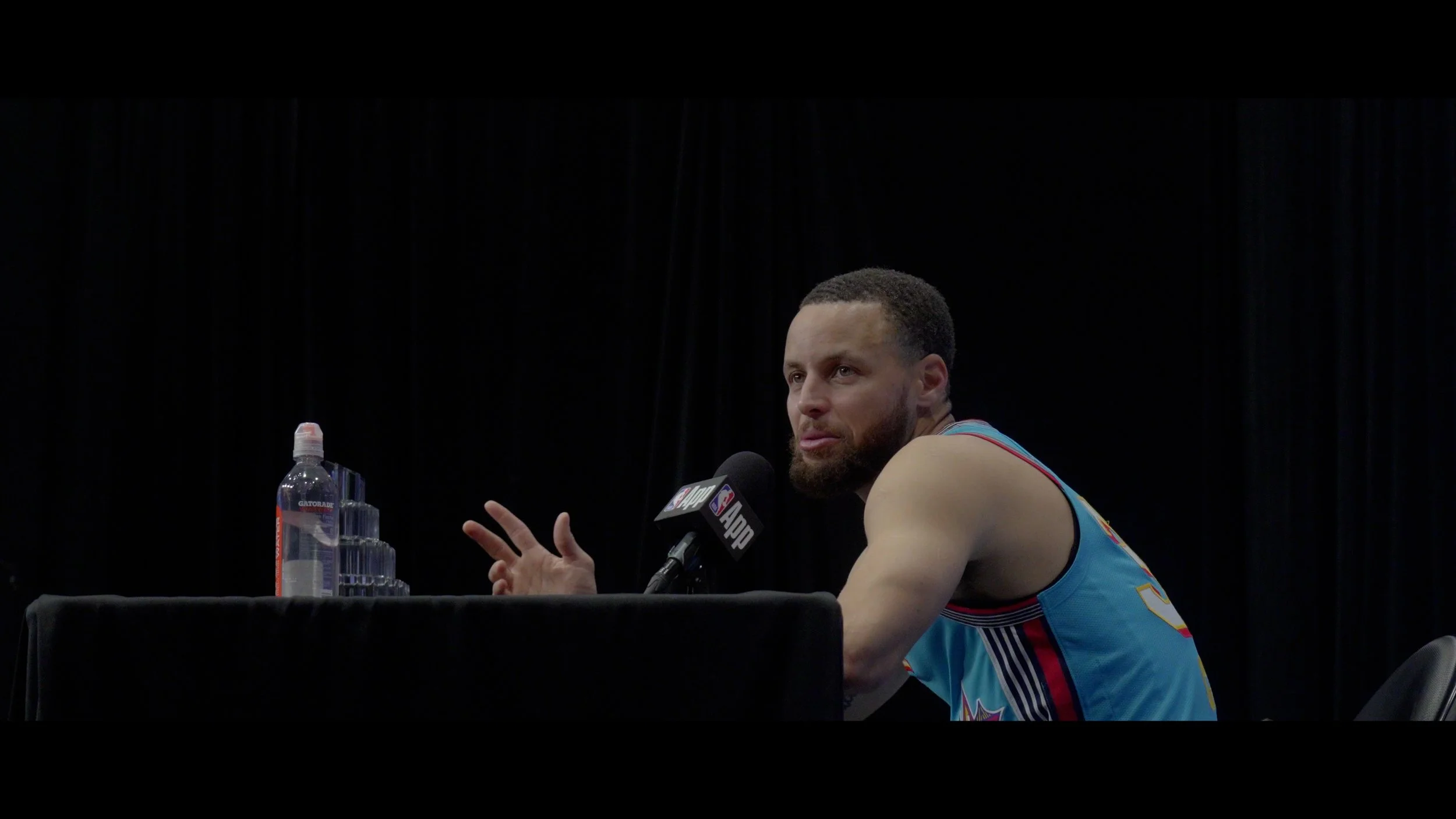A basketball player with a beard and short hair, wearing a teal jersey, speaking into a microphone with the NBA logo, sitting at a table with water bottles in front of him, against a black background.
