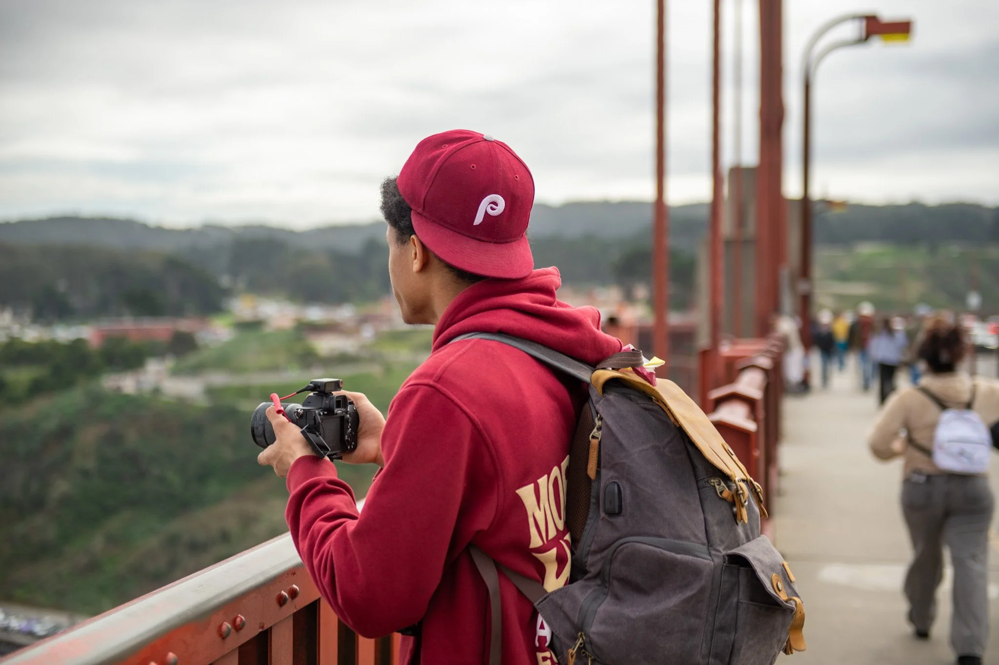 A person wearing a red hoodie and red cap with a P logo, standing on a bridge, holding a camera, and looking at the view of a cityscape with hills in the background.