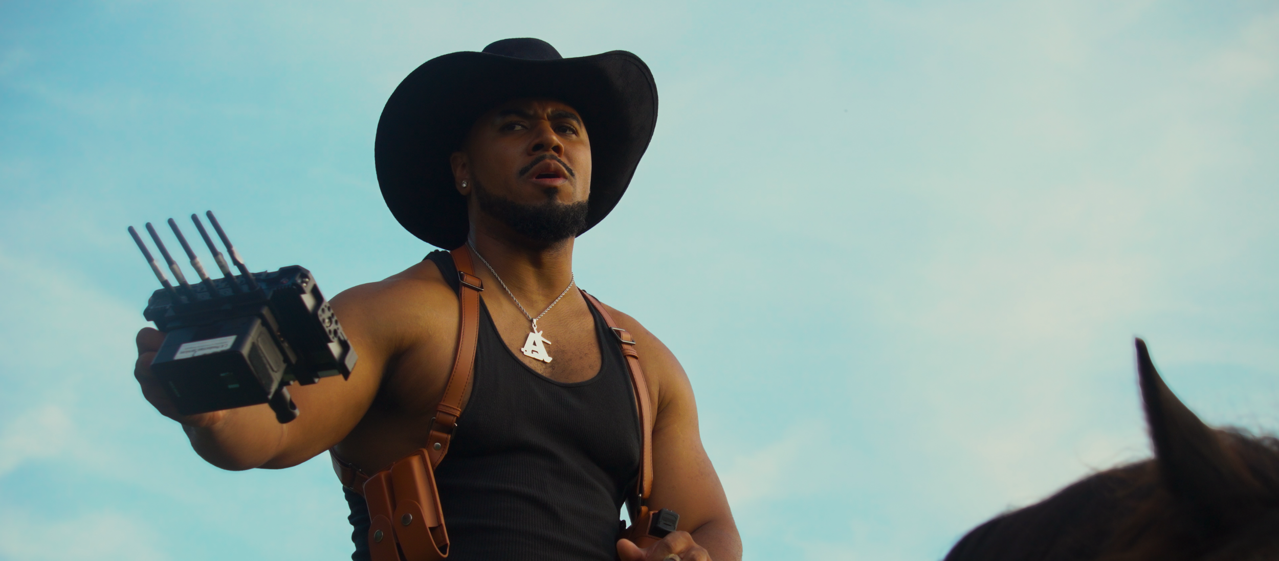 A man wearing a wide-brimmed black hat, a black tank top, and a silver necklace with a pendant, holding a device with multiple antennas, standing outdoors against a blue sky.