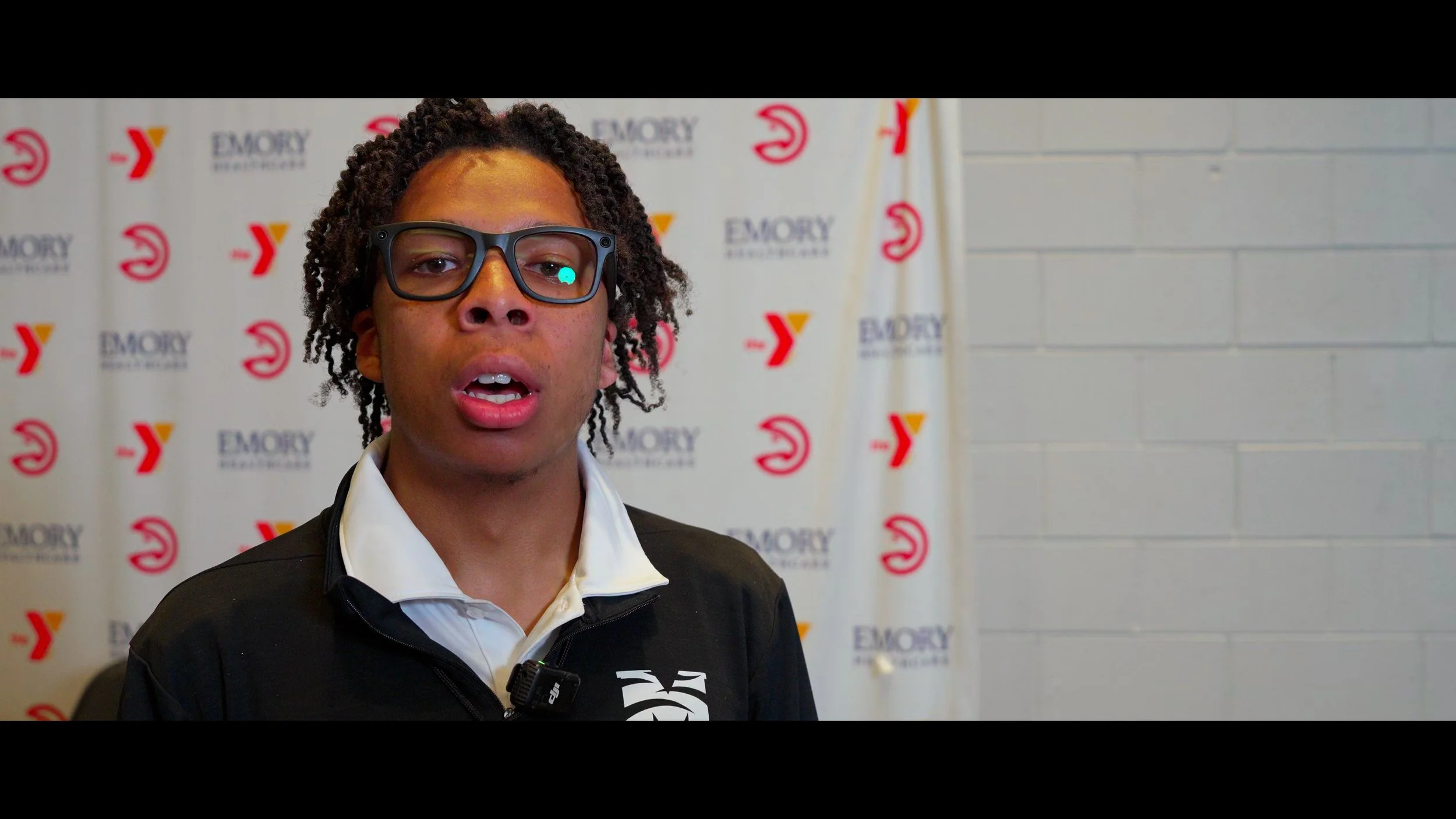 Young man with curly hair and glasses speaking at an event with a backdrop displaying the Emory University Y logo.