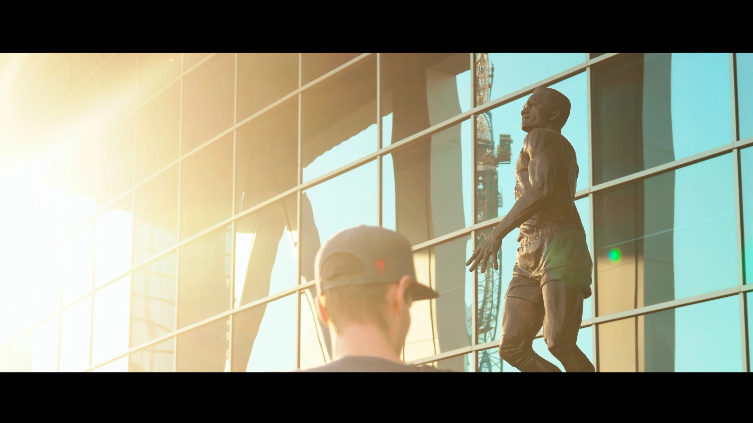 A bronze statue of a man dressed in casual clothes, standing outside a modern glass building, with sunlight reflecting on the glass.