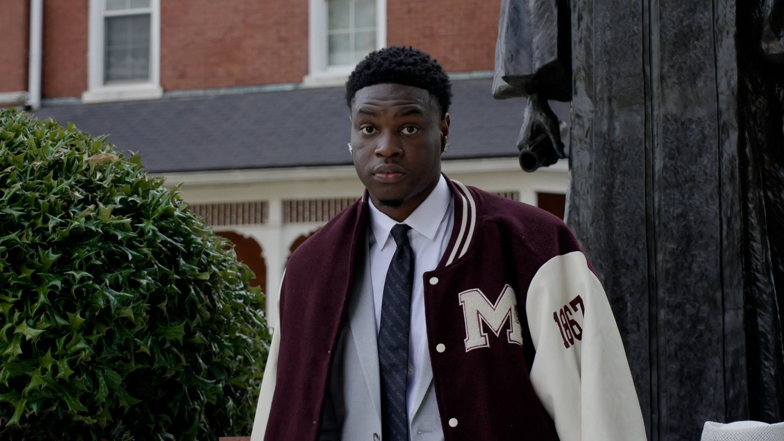 Young man in a maroon and cream varsity jacket with a letter 'M' standing outdoors next to a green bush, with a brick building and windows in the background.