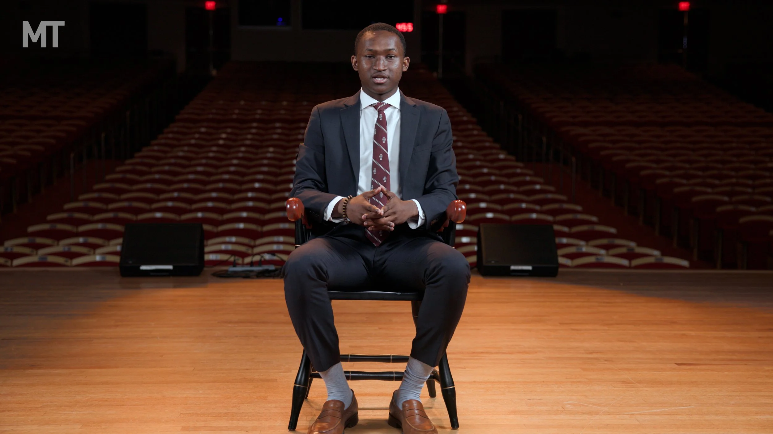 A young man in a dark suit, white shirt, and patterned tie sits on a chair on a stage in an auditorium with empty red and beige seats behind him. He has a dark bracelet on his left wrist and is looking directly at the camera. The stage has wood flooring, and there are two black speakers on the floor beside him.