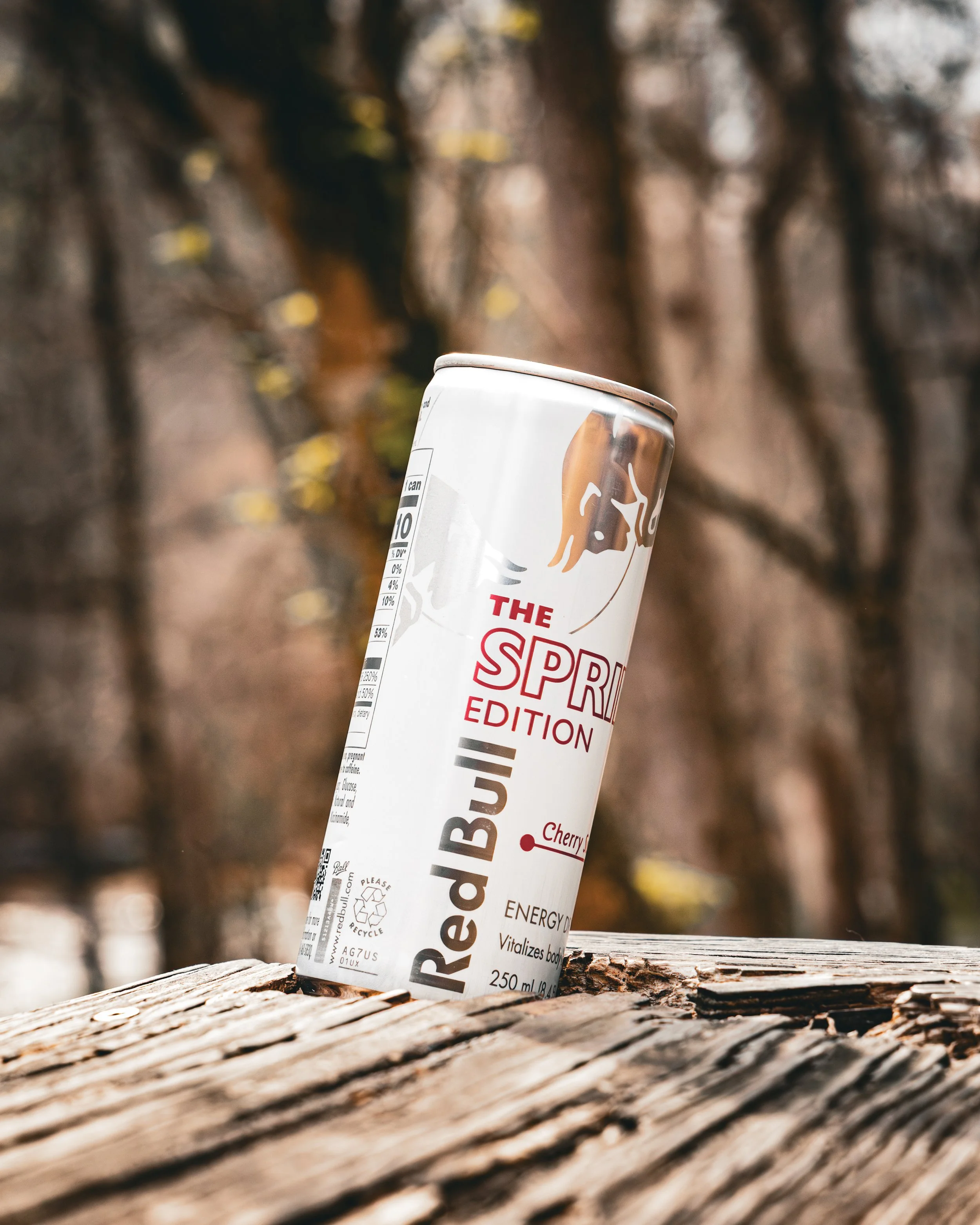 A can of Red Bull energy drink placed on a weathered wooden surface outdoors with a blurred natural background.
