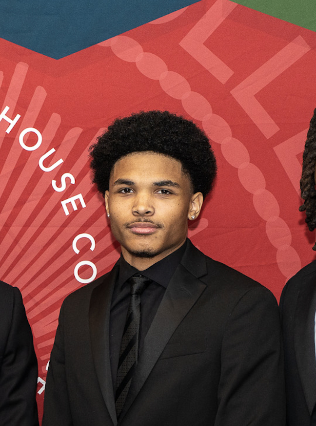 Young man with curly hair in a black suit, black shirt, and black tie posing at an event with a red background.
