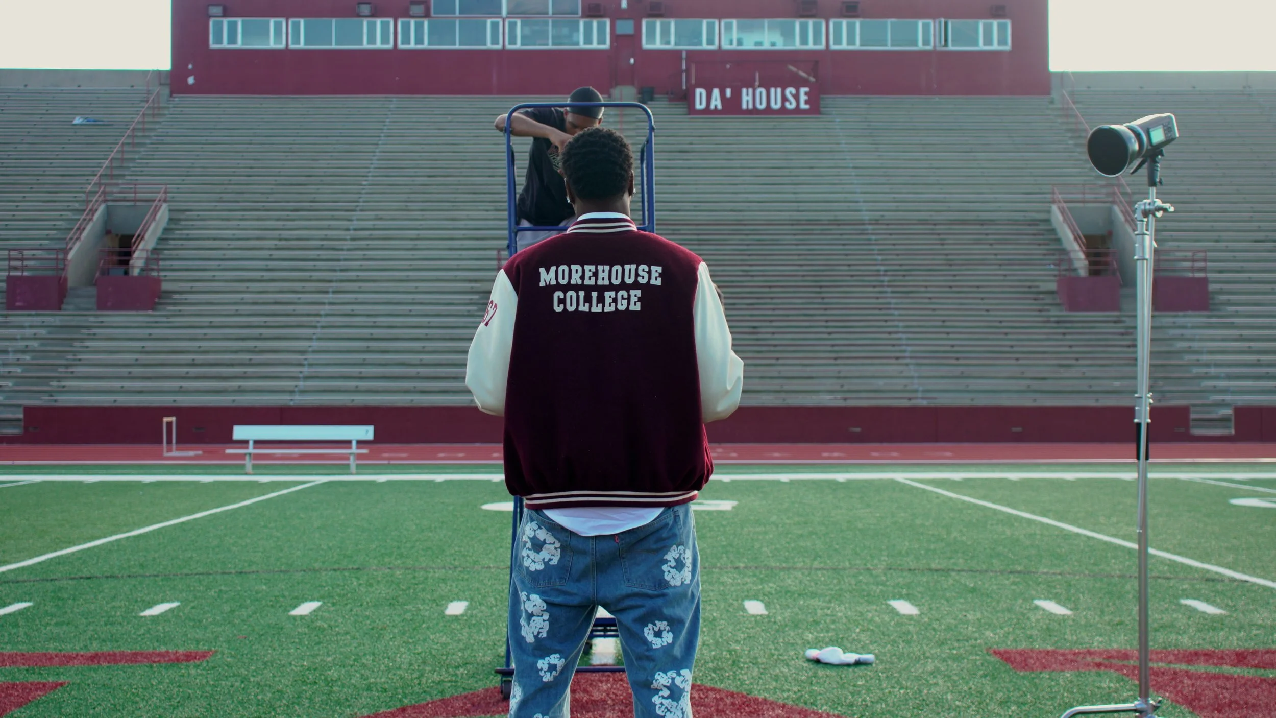 Two young men on a football field, one wearing a jacket that says 'Morehouse College' on the back, practicing with a football passing device. The stadium has empty bleachers and a sign that reads 'DA' HOUSE' in the background.