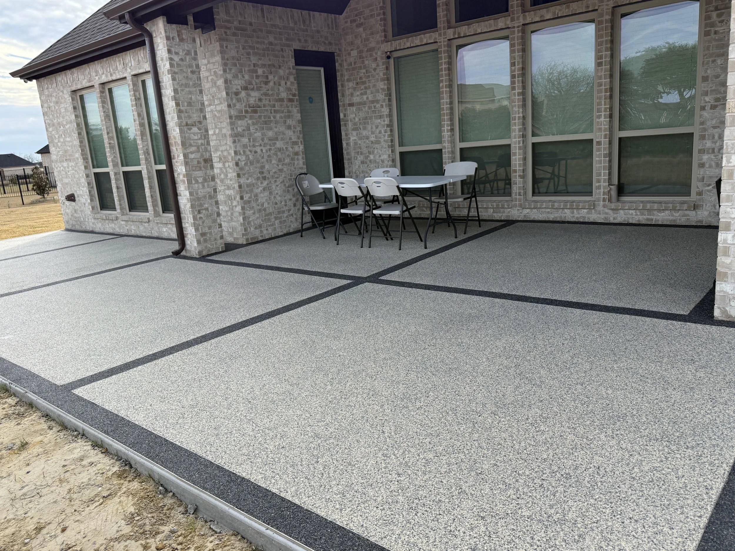A newly finished concrete patio with black border accents outside a brick house. The patio includes a white table with five white and black chairs and has a sliding glass door leading into the house. A black downspout is attached to the house, and a 