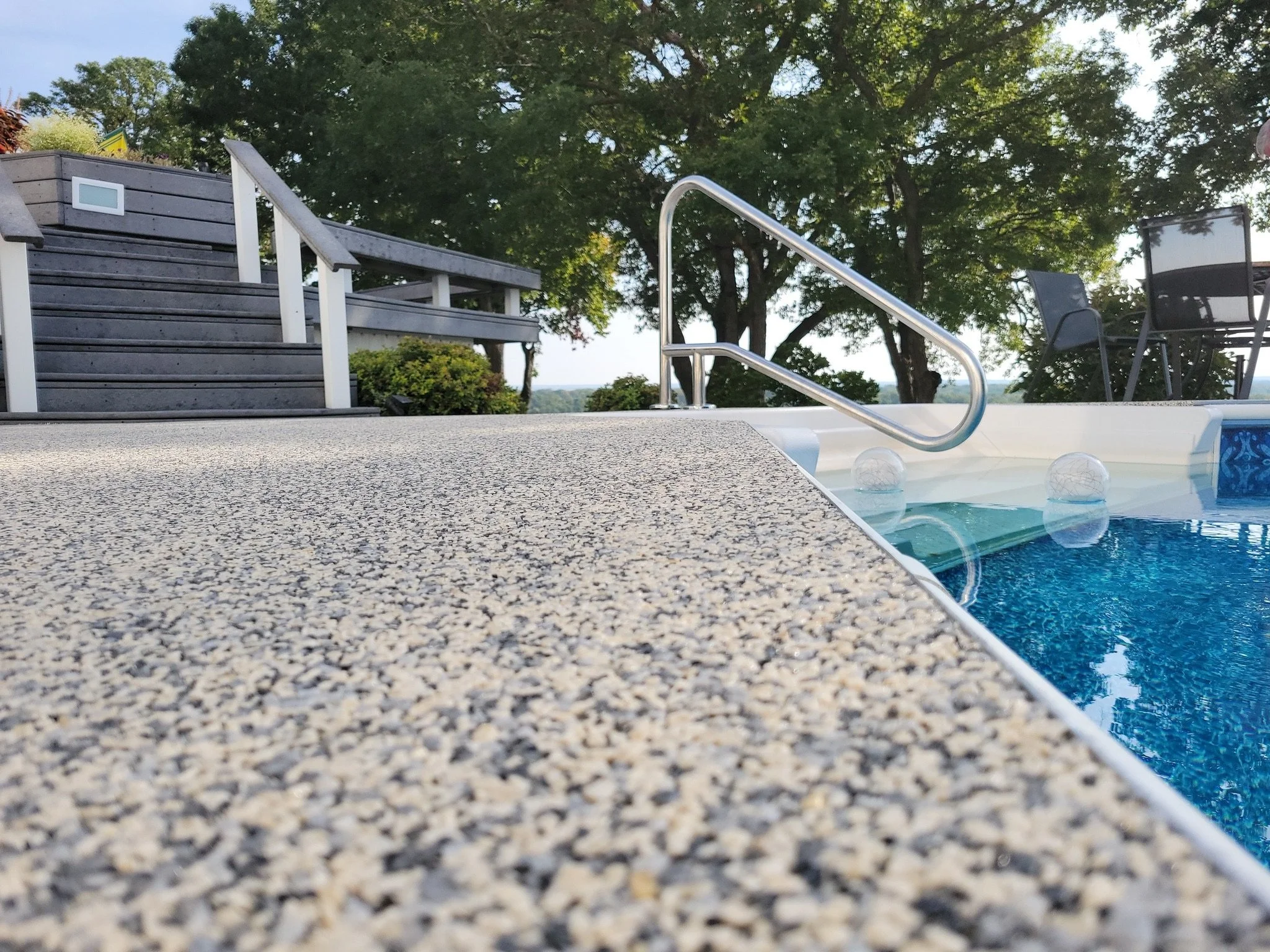 Close-up view of a backyard swimming pool with a textured concrete deck in the foreground, stainless steel handrails leading into the water, and patio furniture including chairs and a table, with trees and clear sky in the background.