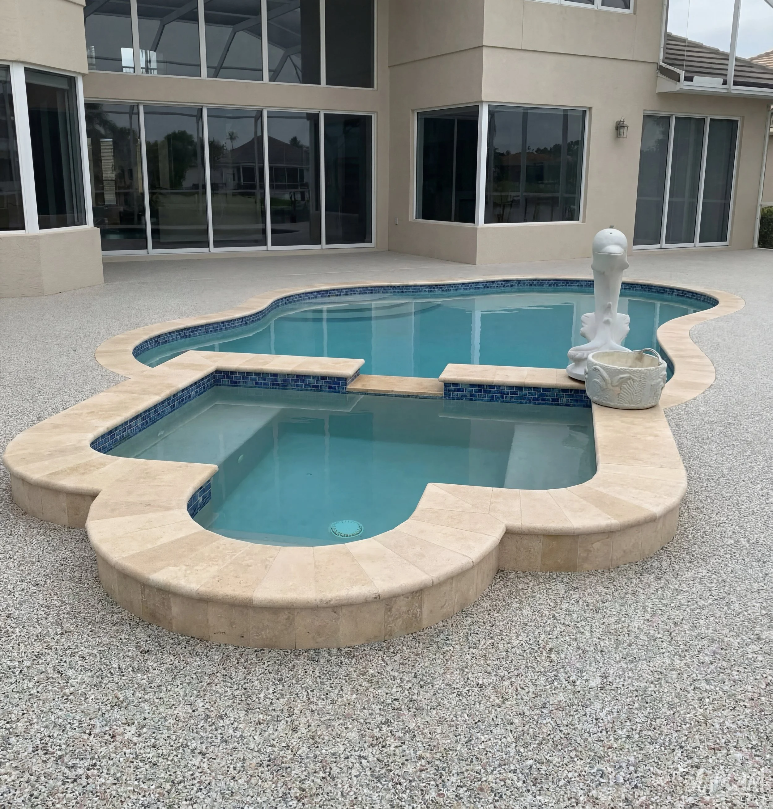 A backyard with a multi-section in-ground pool with beige stone edges, a small fountain, and a white decorative urn, adjacent to a beige house with large windows.
