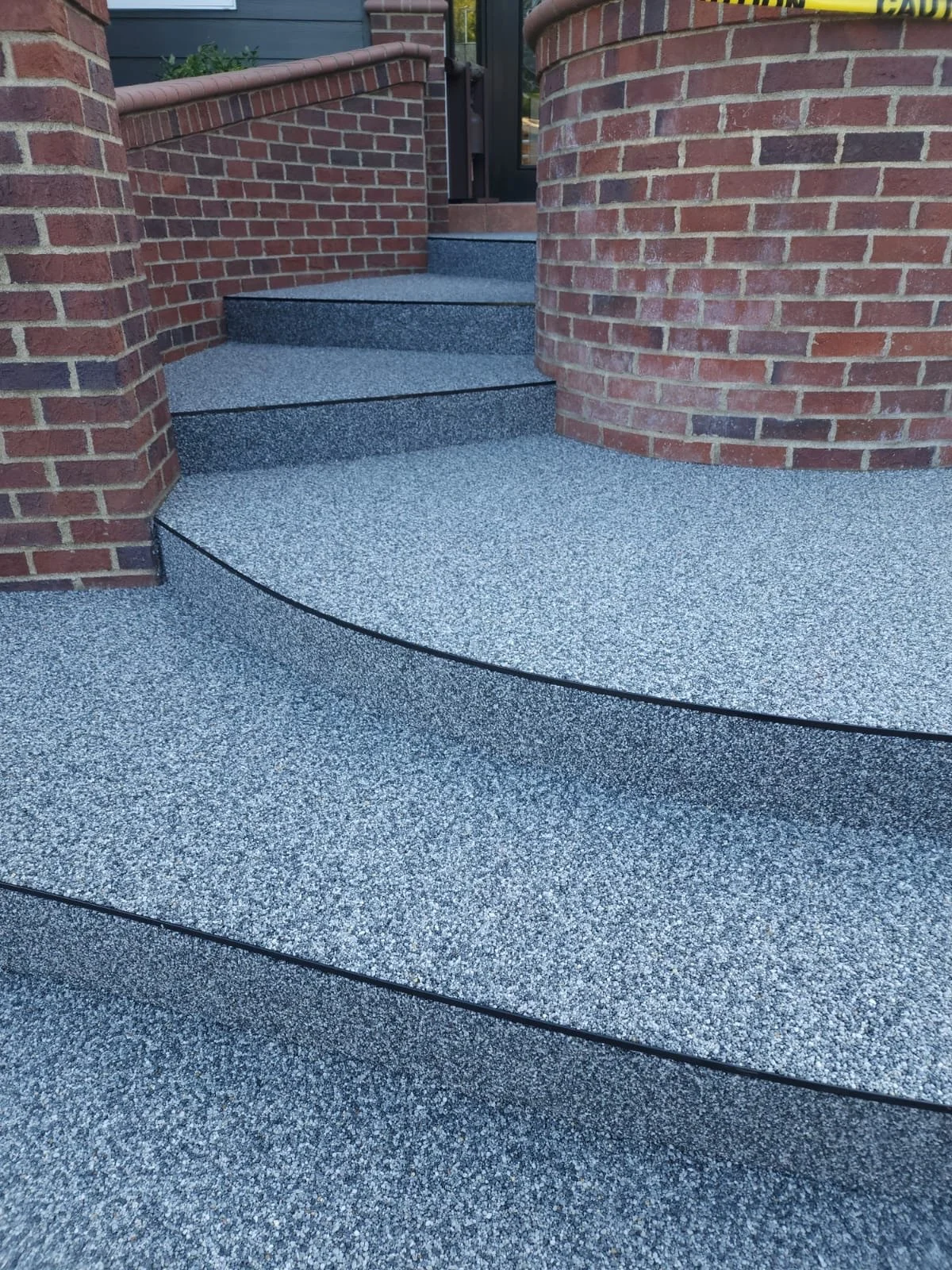 Close-up of textured gray and white speckled outdoor stairs with black edging, leading up to a door, surrounded by red brick walls.