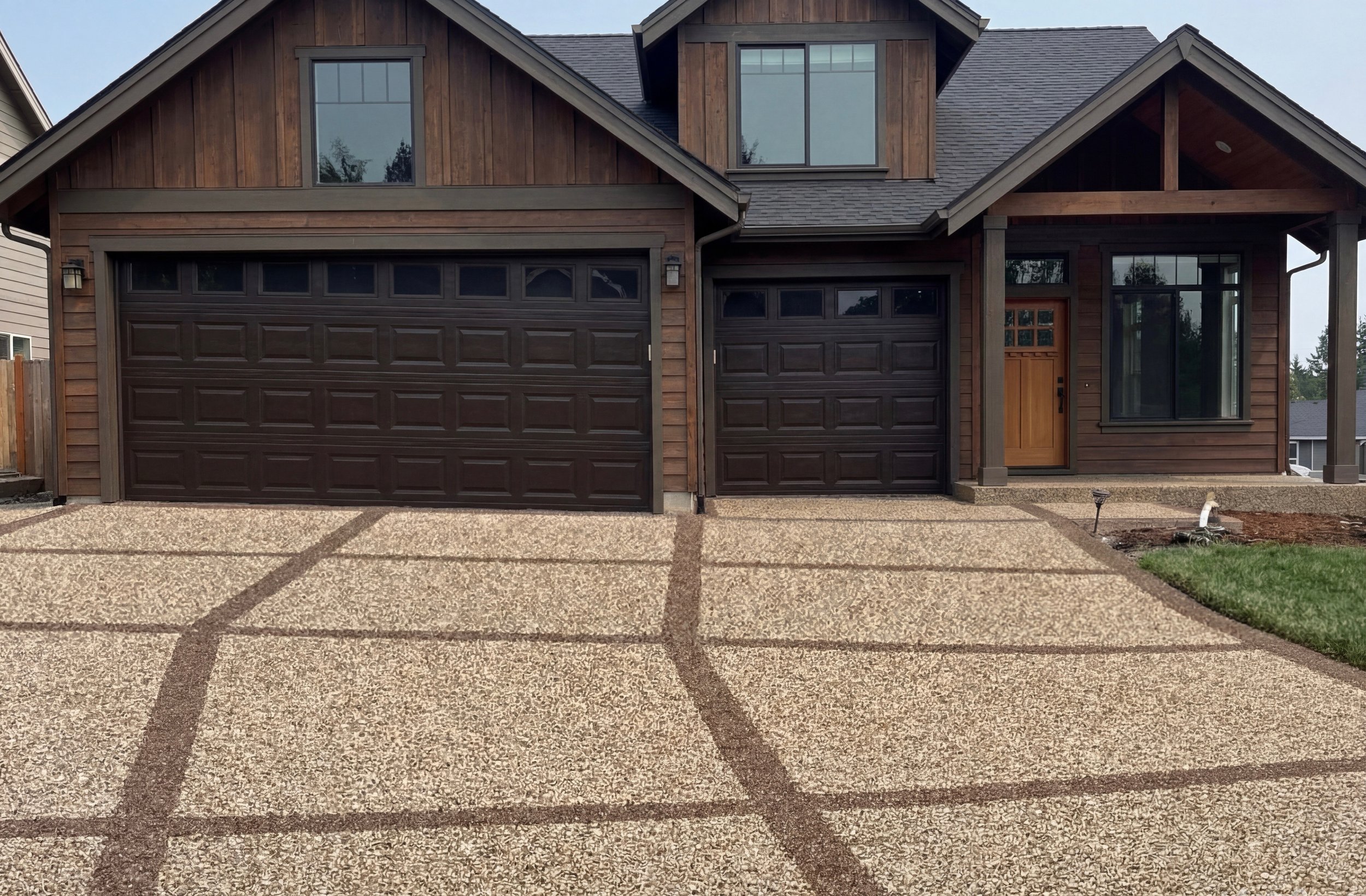 Front view of a modern two-story house with a dark brown garage door, a wooden front door, and large windows. The driveway is made of textured concrete with decorative brown lines. There is a small front lawn and some exposed pipework near the house'