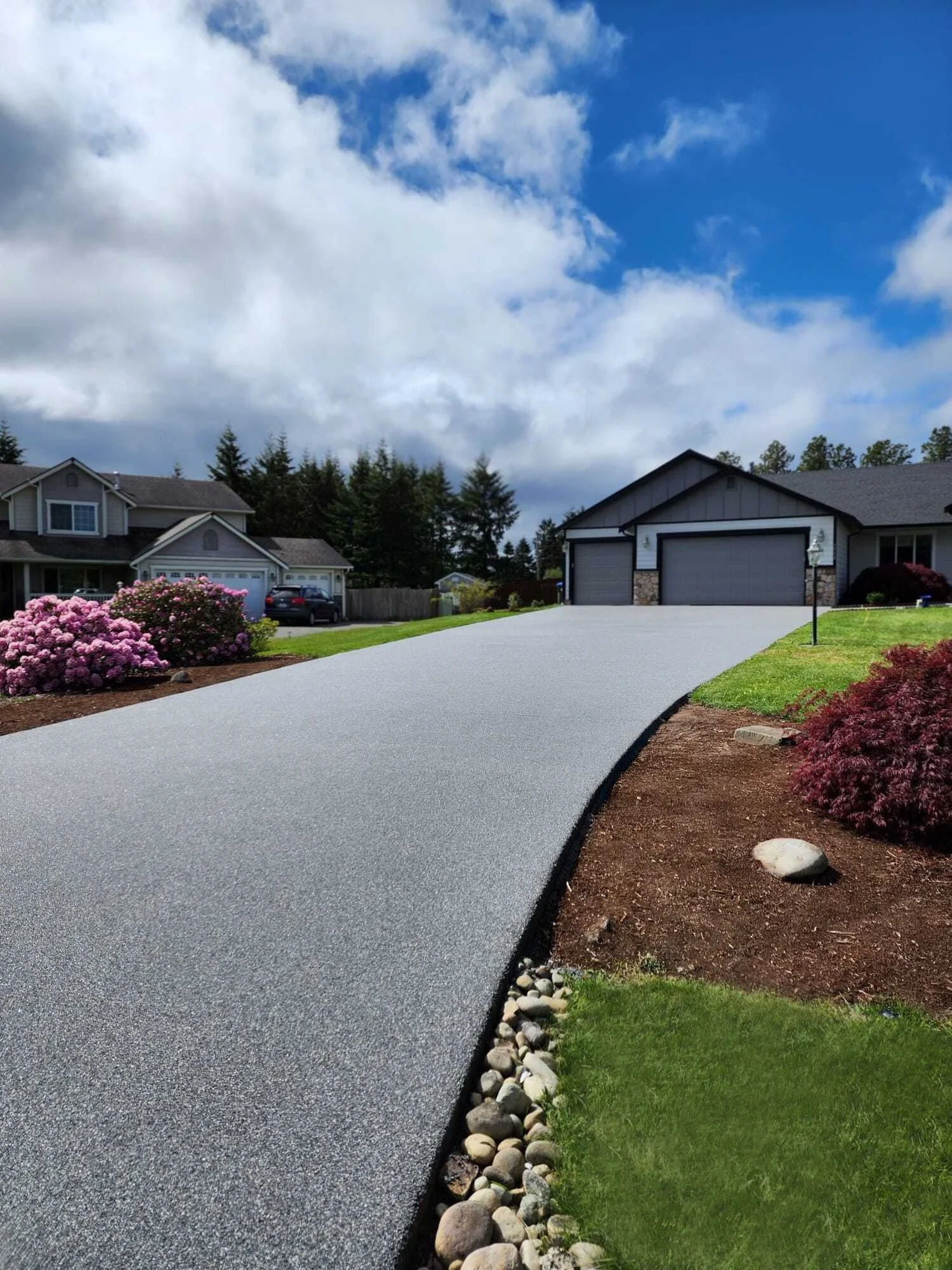A residential driveway leading to a garage, surrounded by landscaped plants and flowers, with a partly cloudy sky overhead.