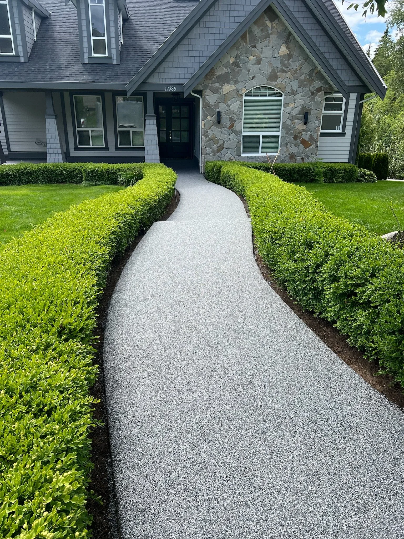 Curved concrete walkway leading to the front door of a house with a stone and siding exterior, surrounded by well-maintained green bushes and lawn.
