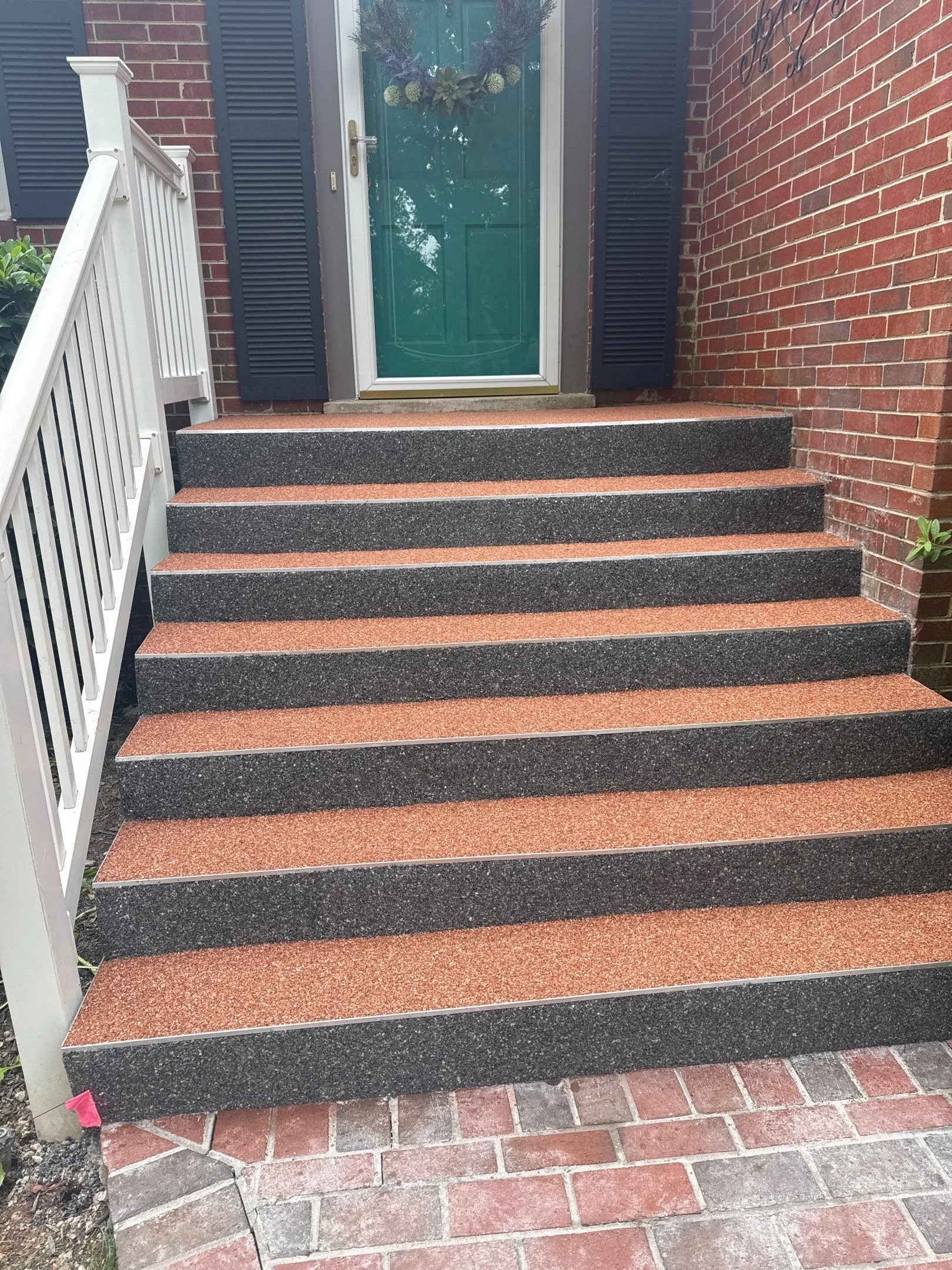 Front porch stairs with pink, black, and red textured steps leading to a teal front door, flanked by black shutters on brick walls, and a white railing on the left side.