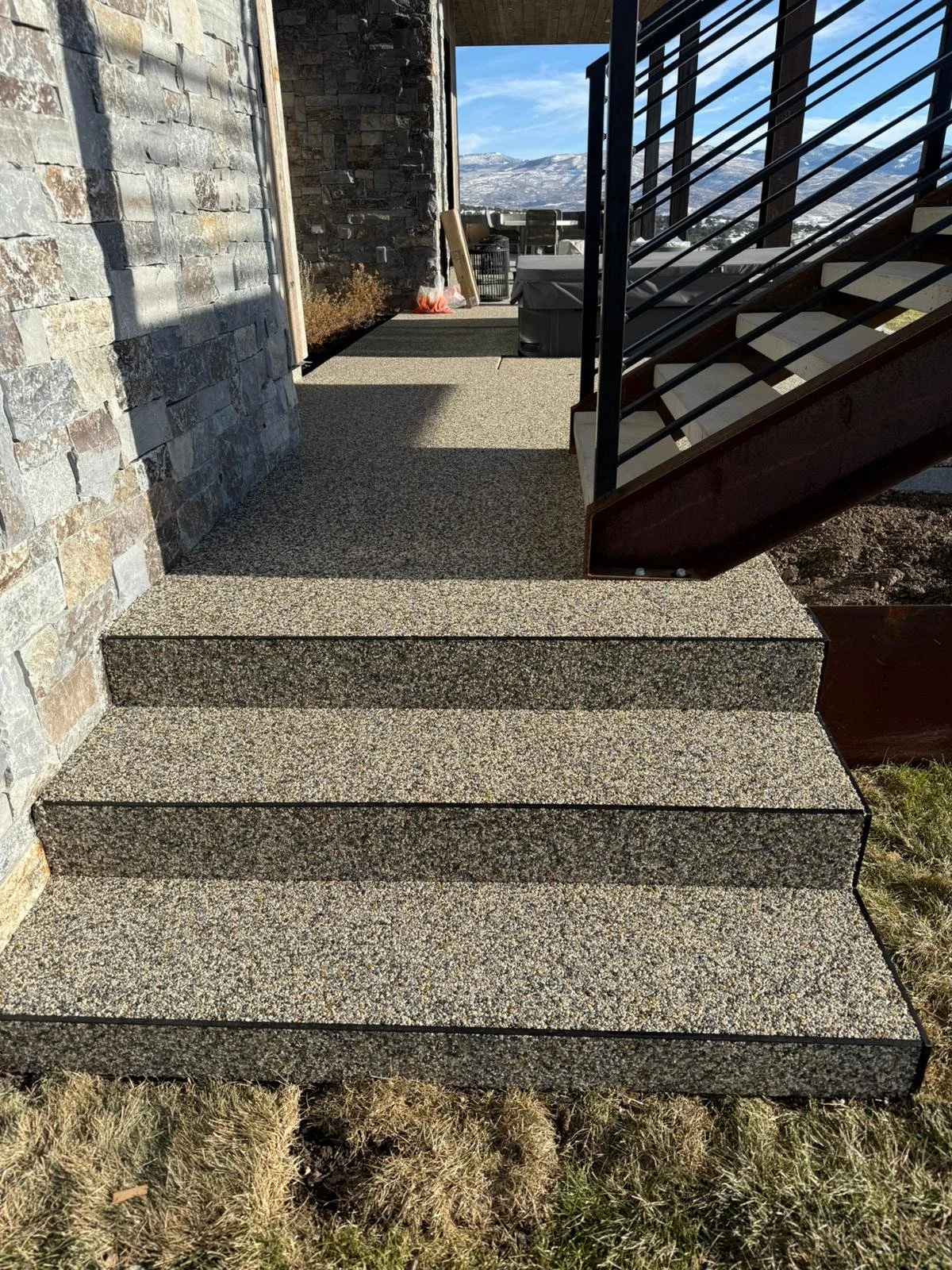 Concrete steps leading up to a patio or balcony area with a stone wall on the left, an outdoor staircase on the right with metal railing, and a mountain view in the background.