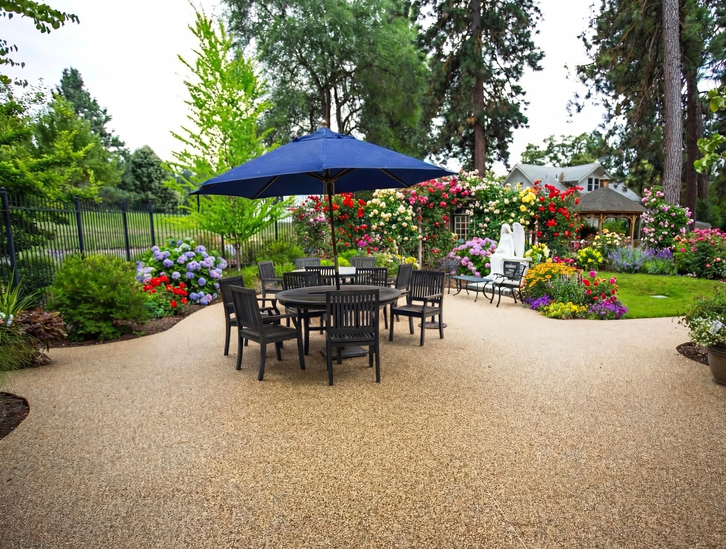 An outdoor garden patio with a round table and chairs under a large blue umbrella. The garden is filled with colorful flowers and trees, with a small white statue and benches in the background.