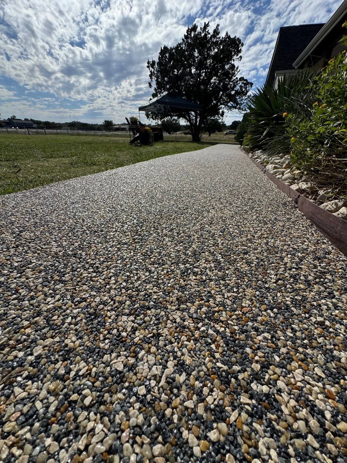 View of a gravel pathway leading to a small outdoor area with a bench, a canopy, and a tree, with a house on the right and a cloudy sky overhead.