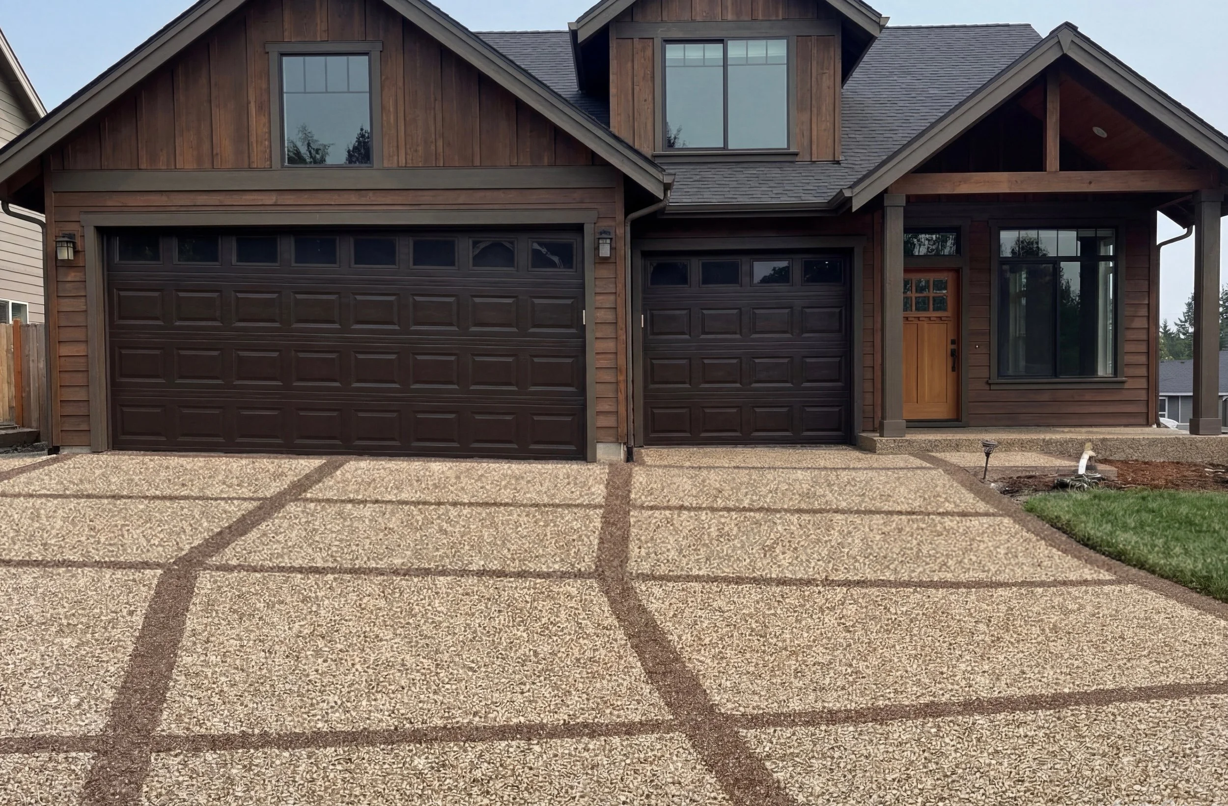 Front view of a modern two-story house with wooden siding, two dark garage doors, a wooden front door, a large front window, and a front yard with a concrete driveway and some grass to the side.