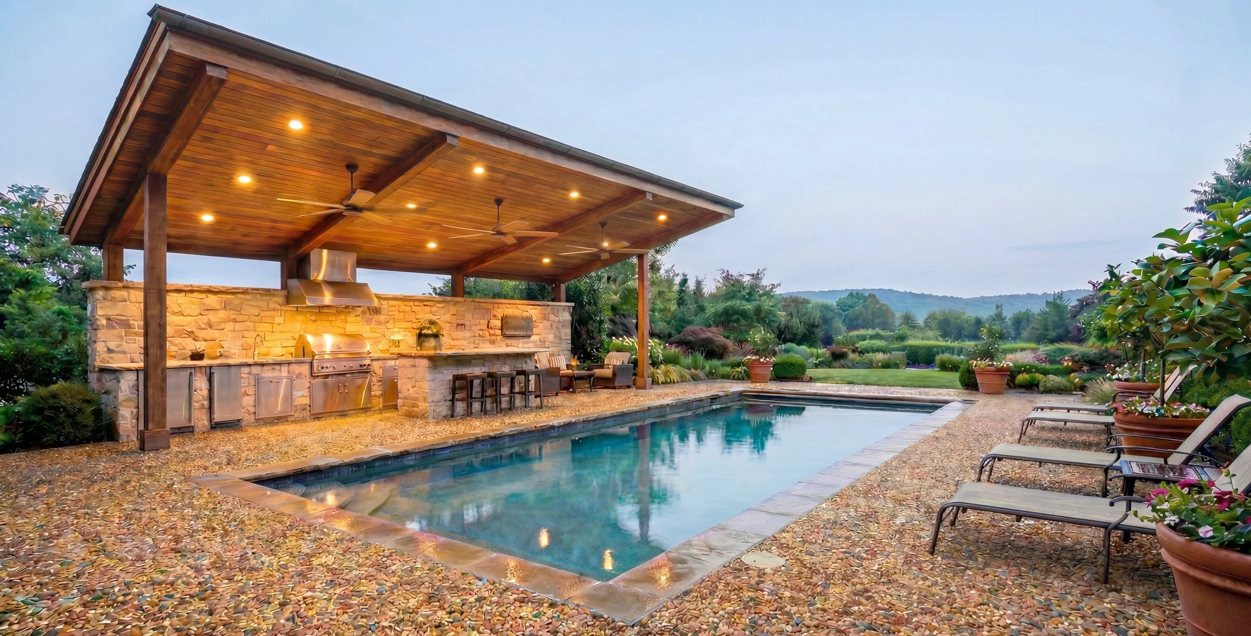 Outdoor backyard with swimming pool, covered outdoor kitchen with seating, outdoor lighting, and lush greenery with trees and hills in the background.