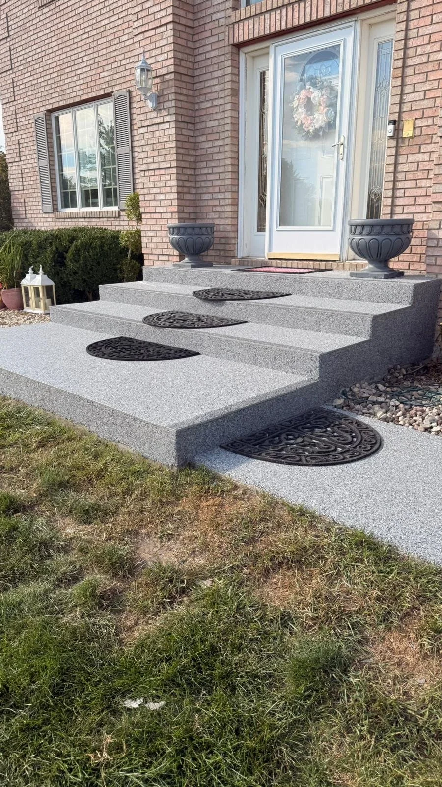 Front porch with gray concrete steps, black decorative doormats, potted plants, and a brick house with a white door adorned with a wreath.