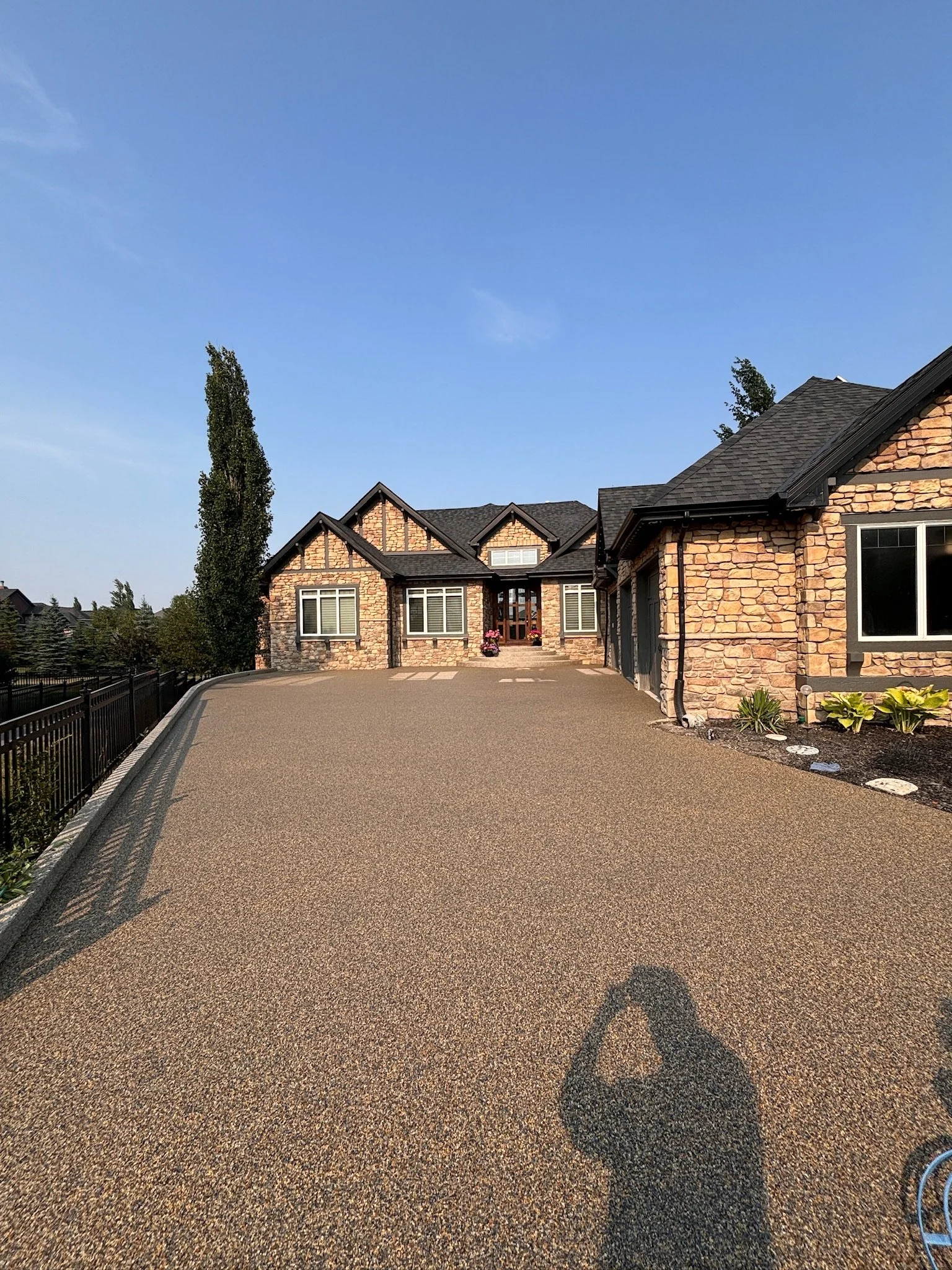 A large house with stone exterior walls and a sloped roof, situated on a driveway with a gravel or asphalt surface. The house has multiple windows and a front porch with potted plants, with trees and landscaping along the edges of the property.
