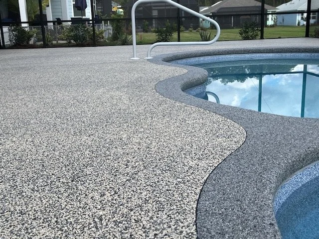 Close-up view of a textured concrete pool deck with a curving edge and a swimming pool with a metal handrail in the background.